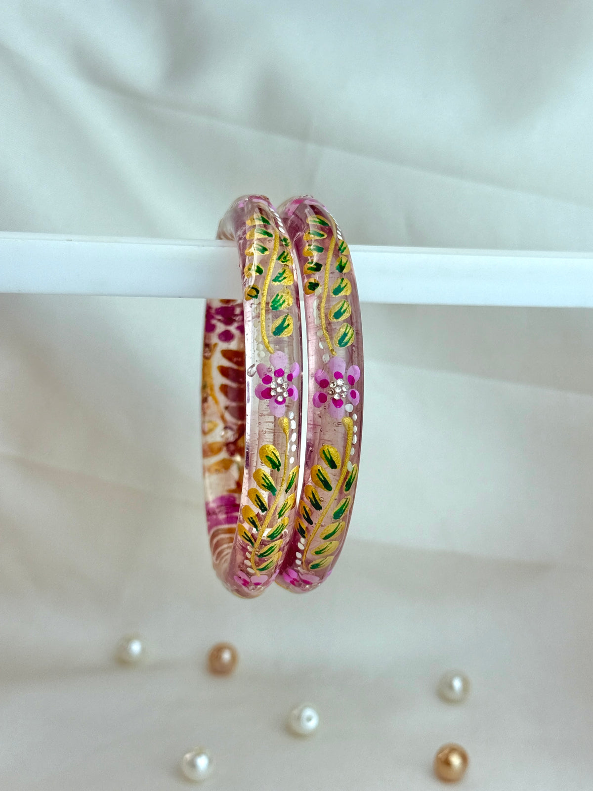 Two pink bangles with colorful floral and leaf patterns, hanging from a white bar against a light fabric backdrop with a few scattered pearl-like beads below.