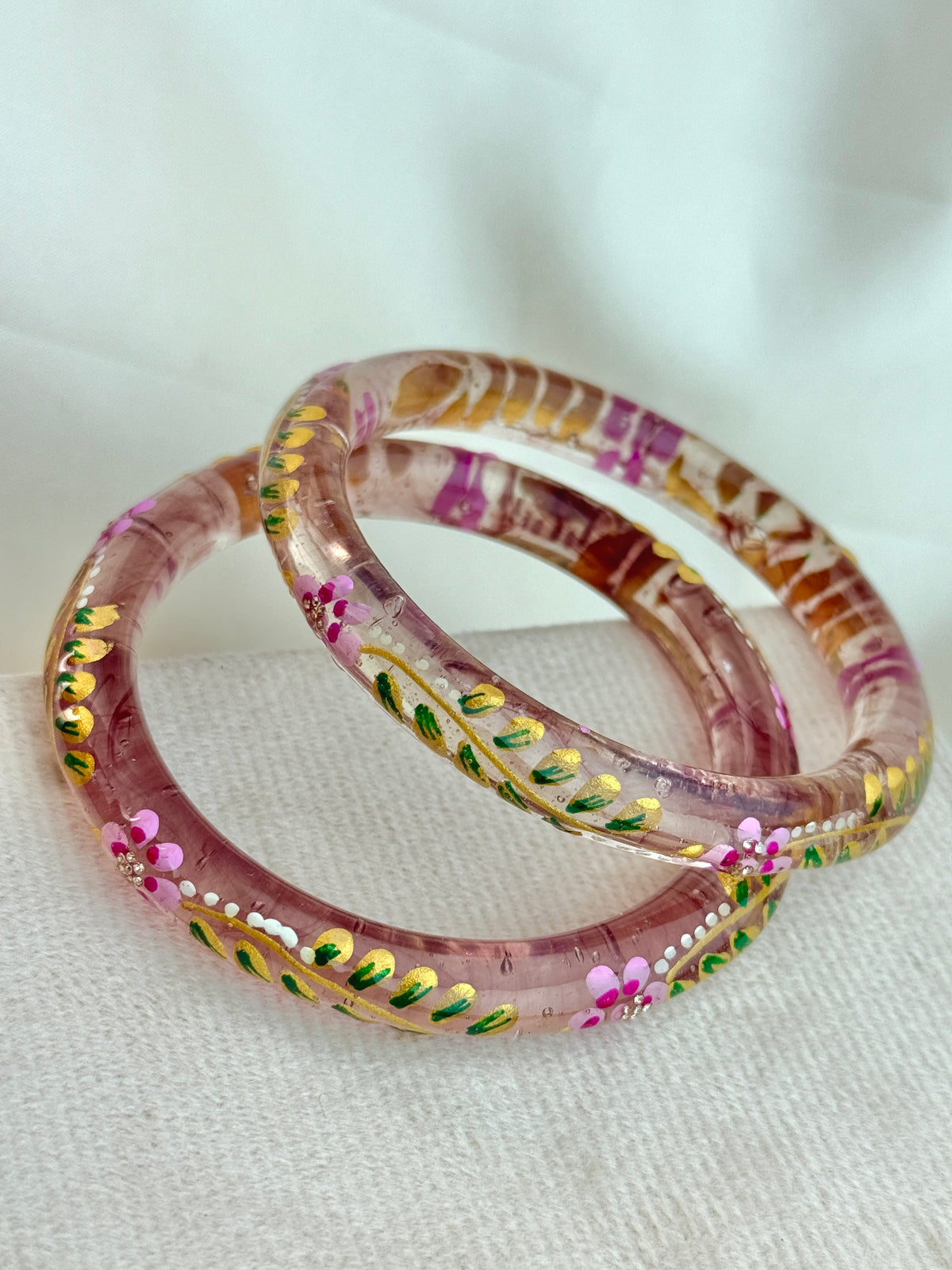 Two pink translucent bangles with hand-painted floral and leaf patterns in pink, green, yellow, and white, resting on a textured white fabric background.