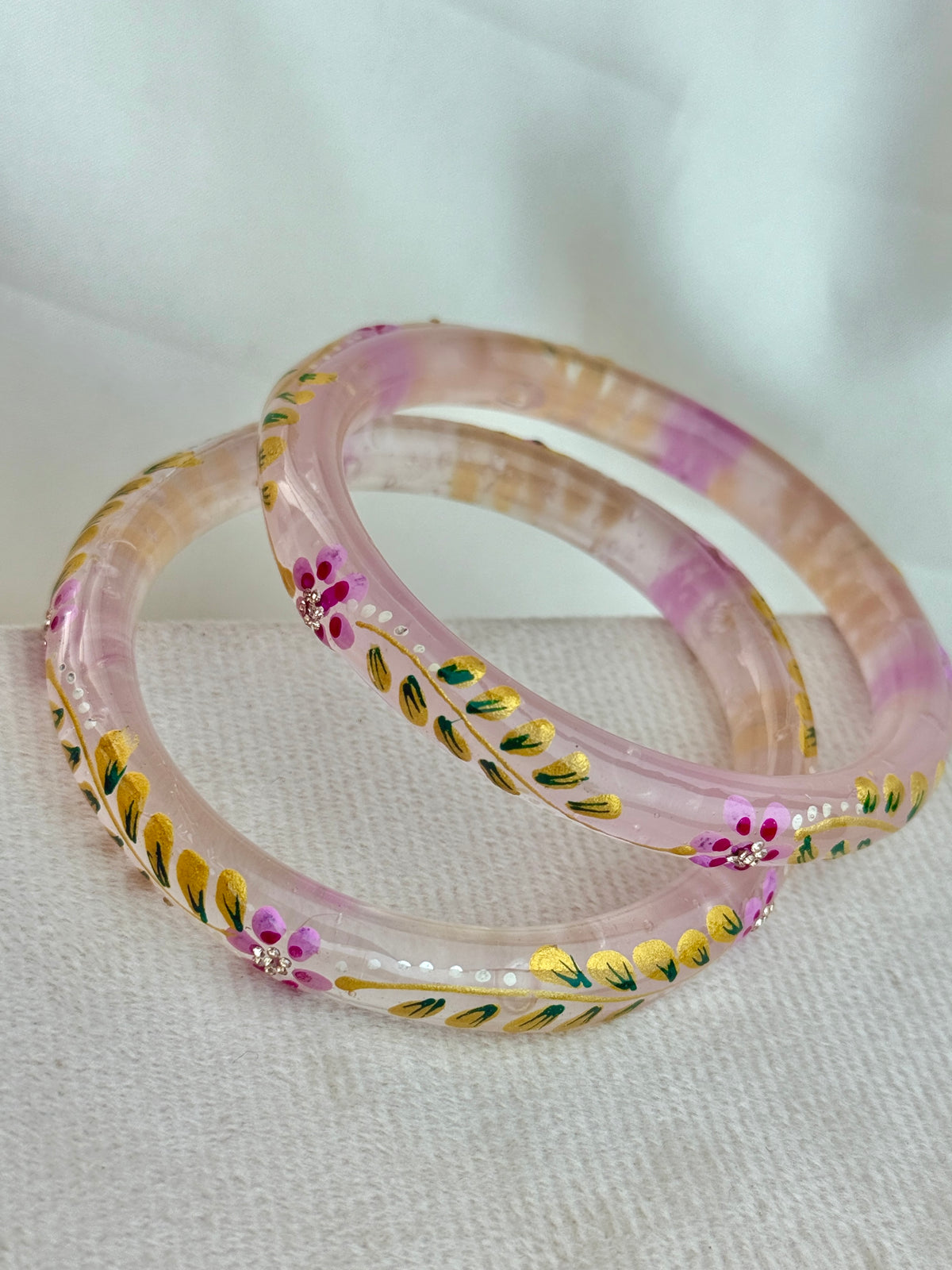 Two pink translucent bangles with hand-painted floral and leaf patterns in yellow, green, and pink, resting on a textured white fabric surface.