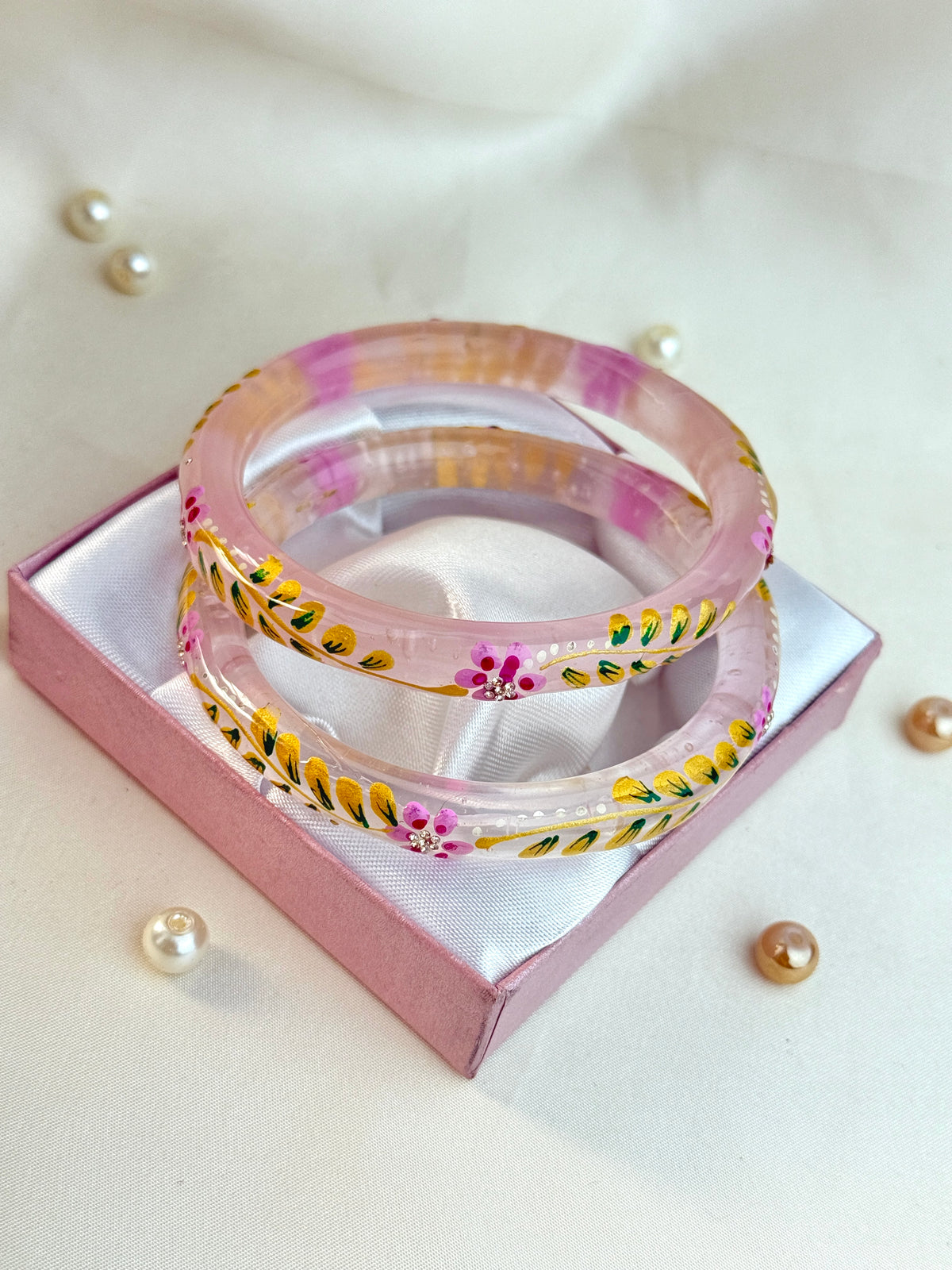 Two pink translucent bangles with colorful floral and leaf patterns, displayed on a pink gift box with a white satin lining, surrounded by small decorative pearls on a light beige fabric background.