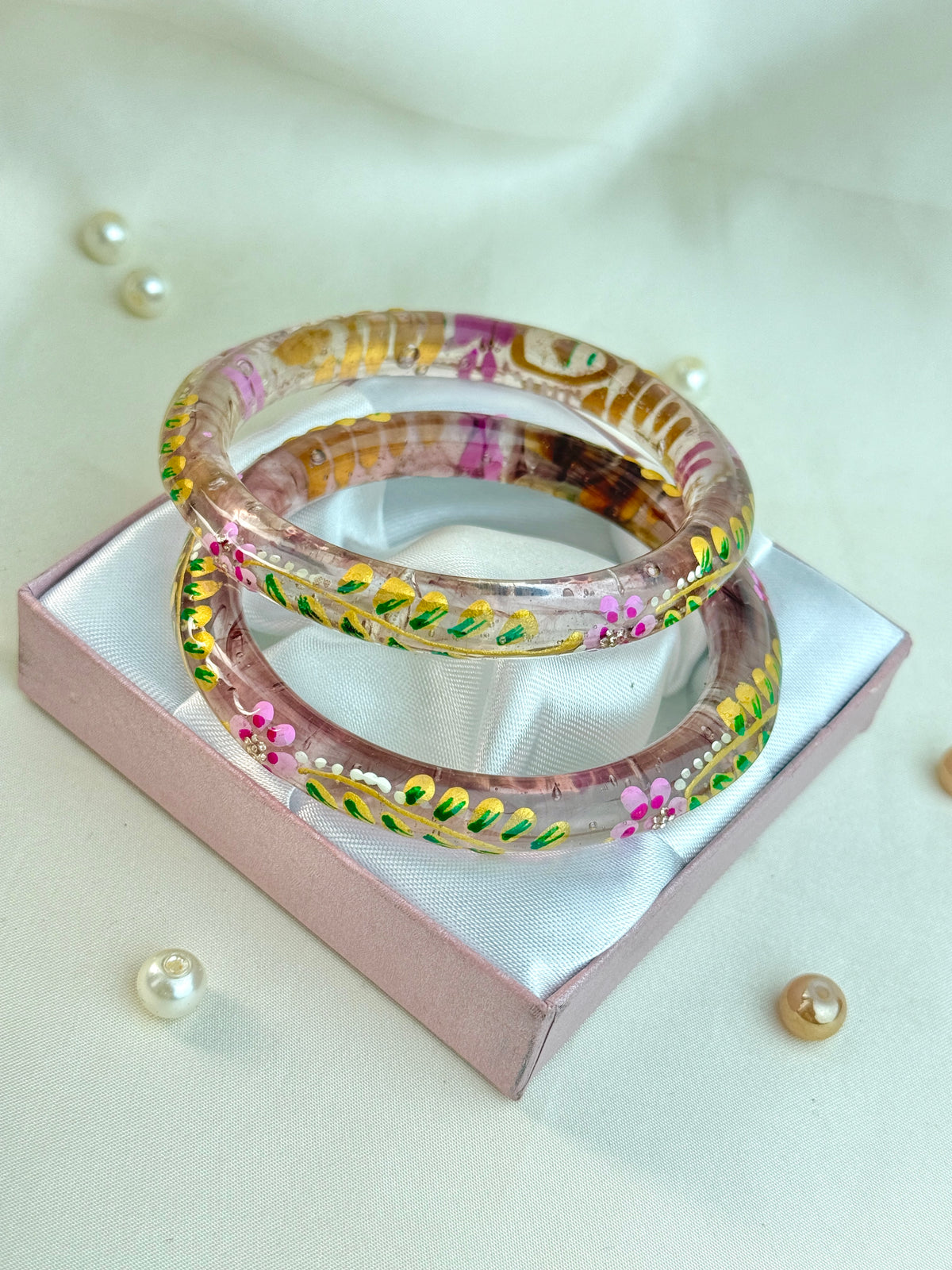 Two stacked clear resin bangles with colorful floral and leaf patterns, displayed on a pink satin-lined gift box with decorative pearls on a light beige fabric background.