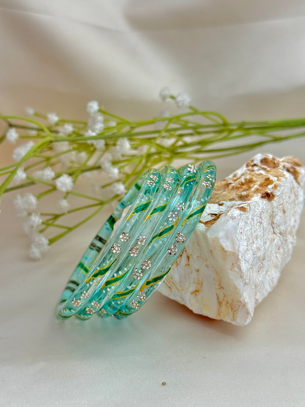 Three translucent aqua glass bangles with green and yellow swirls and small clear rhinestone floral motifs, styled on a cream fabric surface beside white baby's breath flowers and a rough white stone.