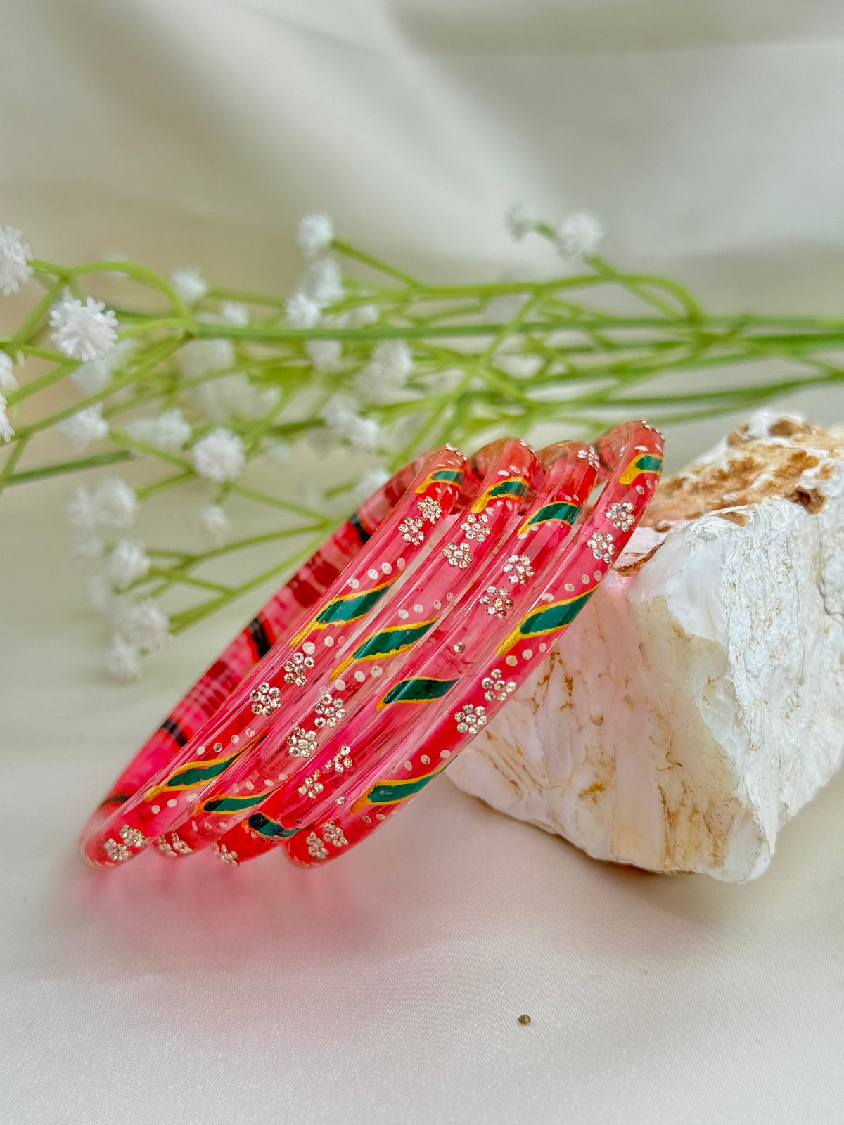 A set of red bangles decorated with white floral patterns and green and gold stripes, resting on a white stone next to baby's breath flowers.