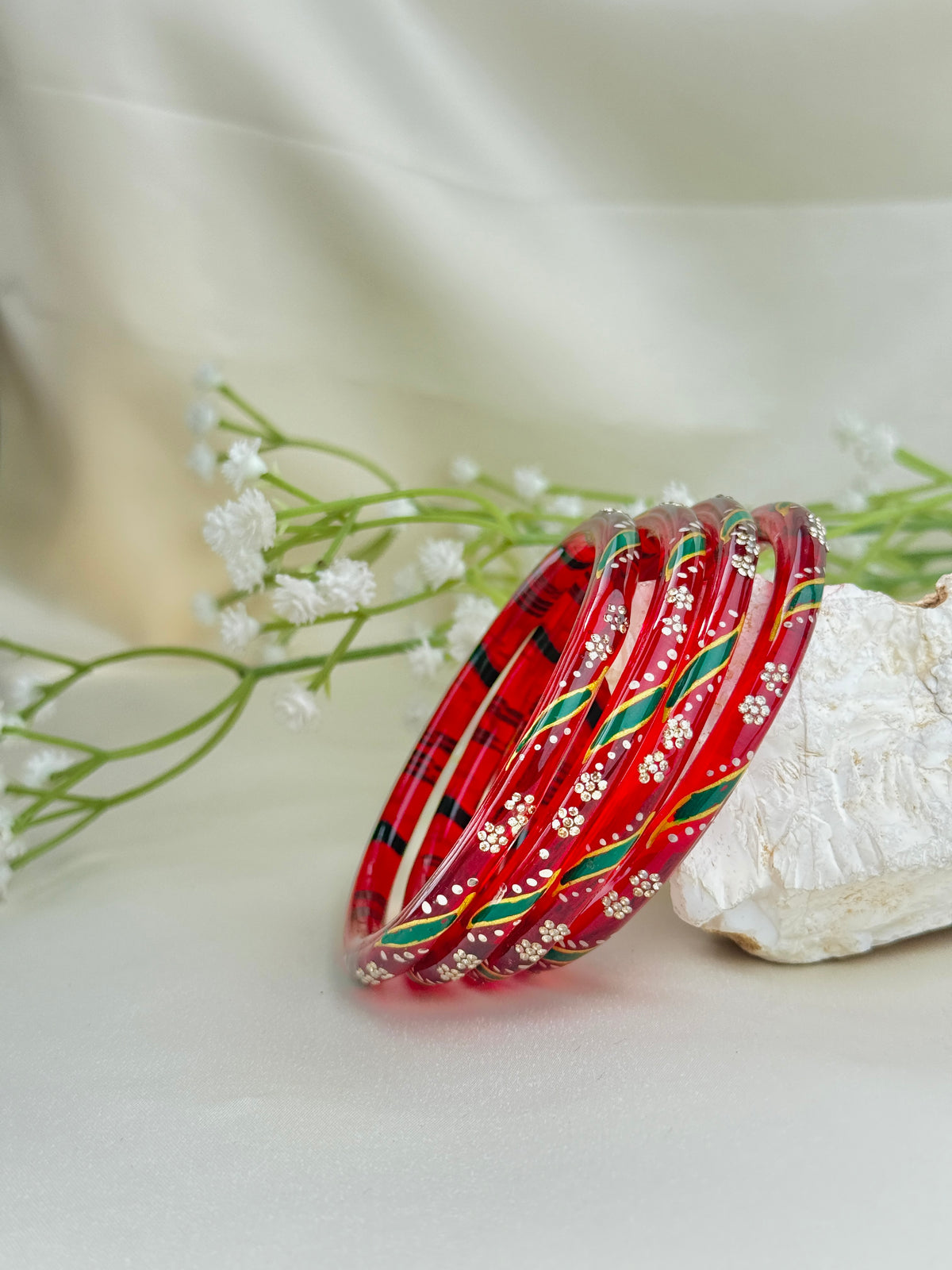 A set of red bangles with white floral patterns and green and black stripes, displayed on a white textured stone with baby's breath flowers against a soft cream fabric background.