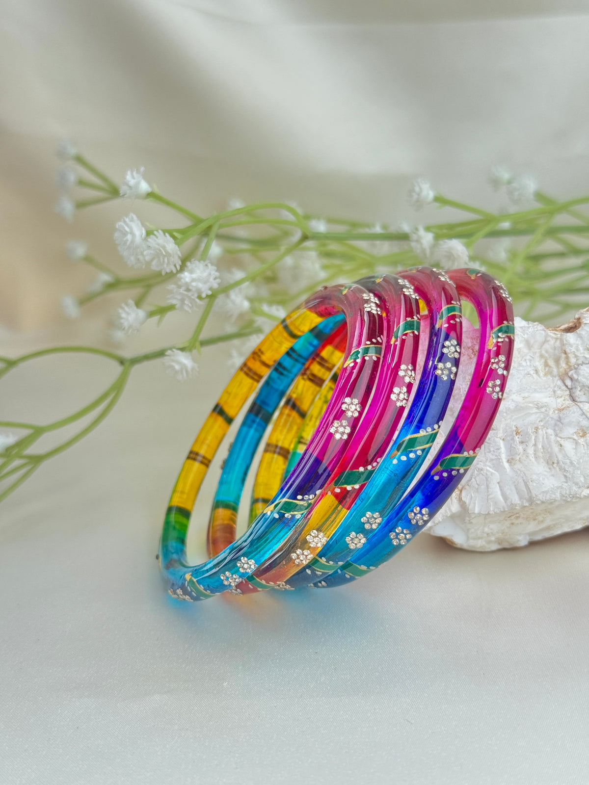 A set of colorful bangles with floral patterns, displayed on a white surface next to small white flowers and a piece of wood.