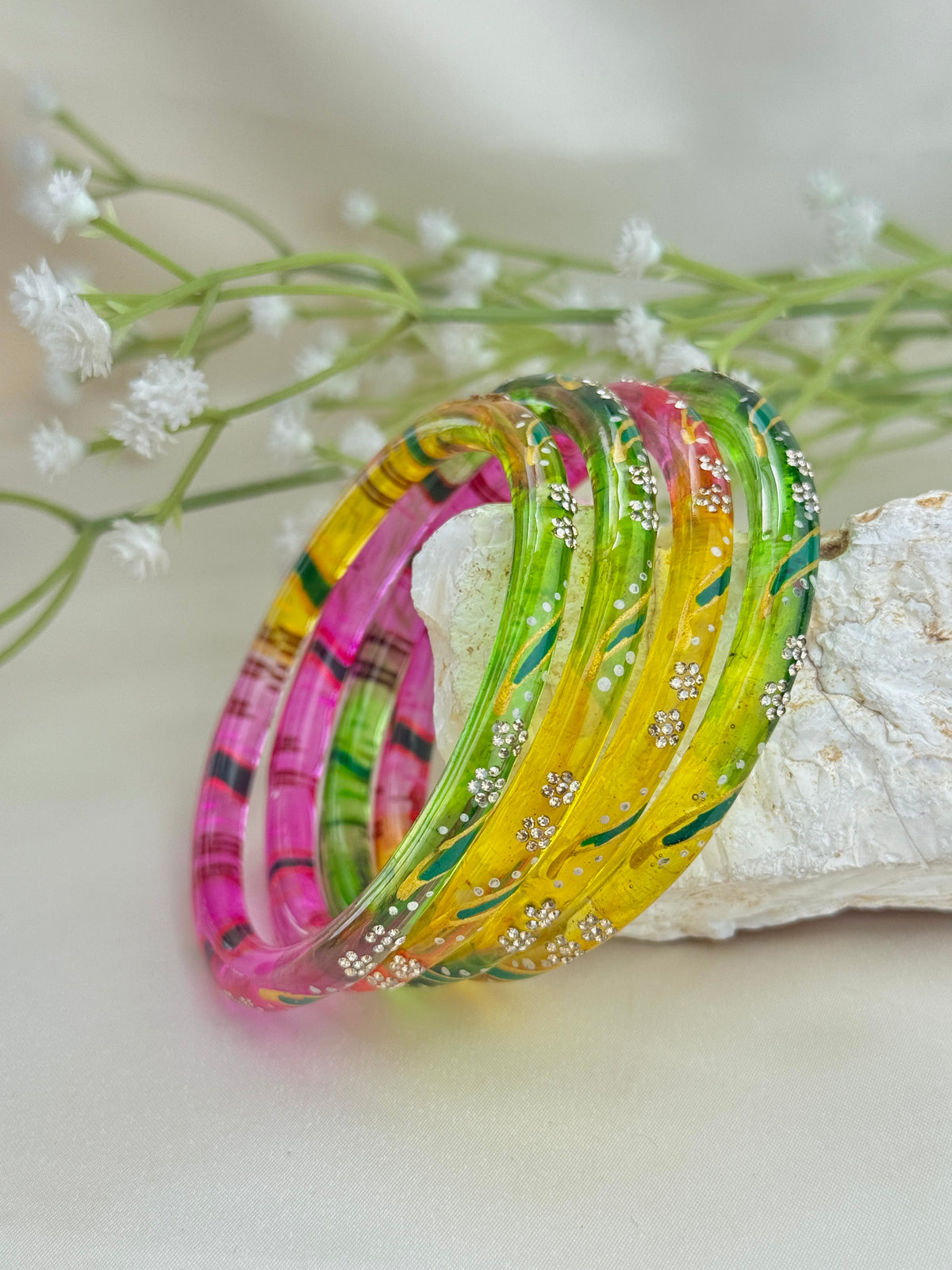 A set of five colorful glass bangles with silver floral details, resting on a white textured stone with delicate white flowers in the background.