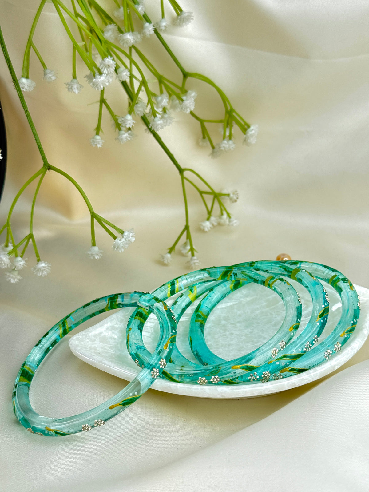 A set of translucent aqua and green marbled bangles with tiny white floral details, displayed on a small white dish with dried baby's breath flowers against a cream satin background.