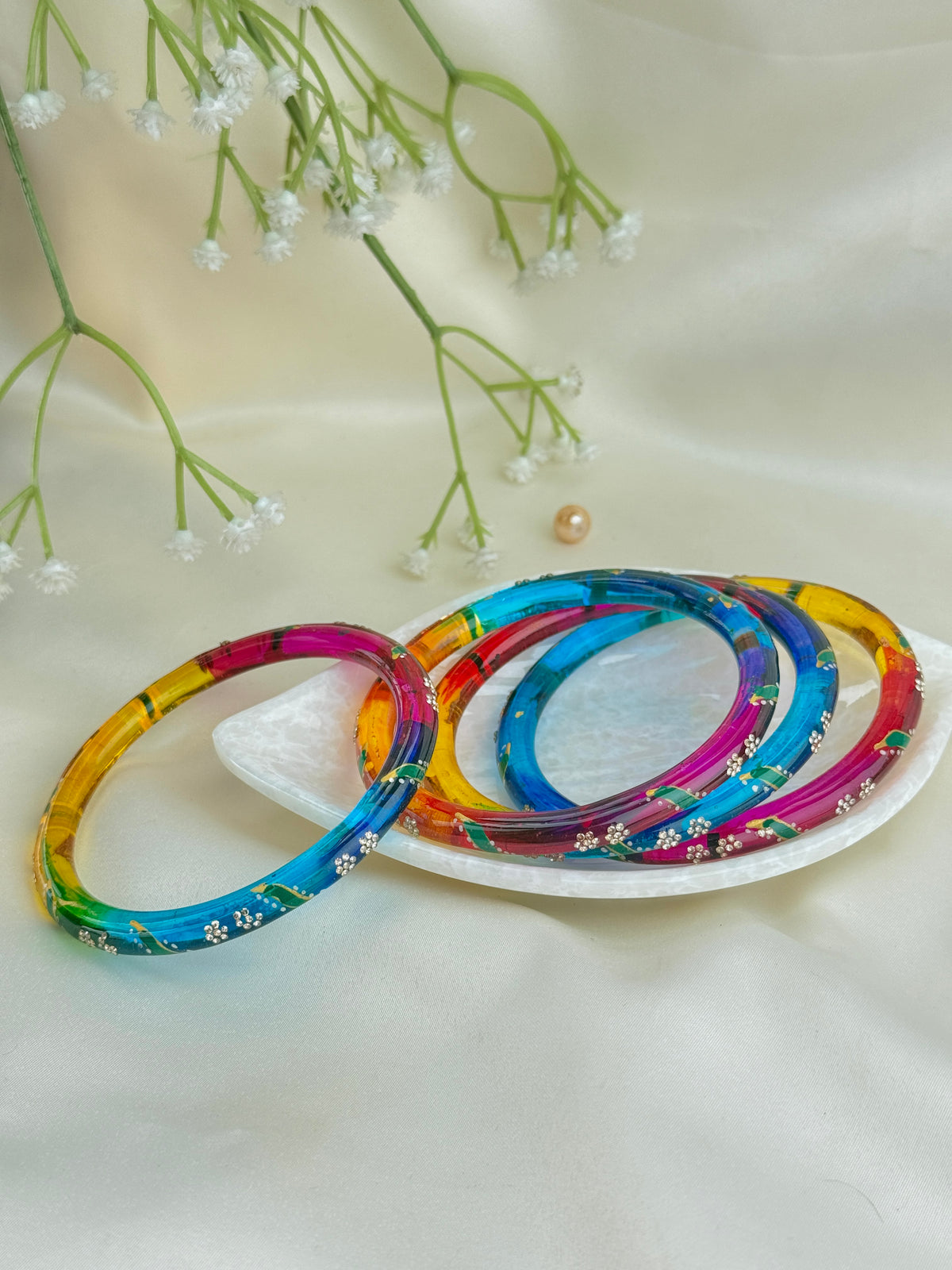 Four colorful acrylic bangles with floral patterns arranged on a small white dish against a cream satin fabric backdrop with baby's breath flowers.
