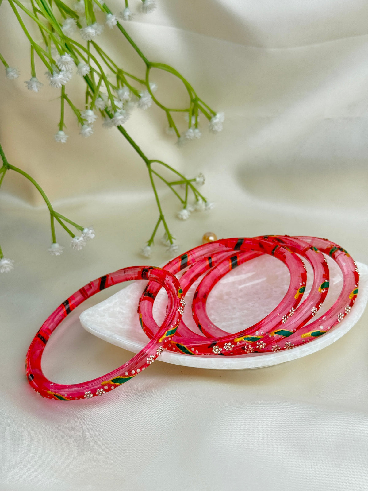 A set of pink bangles with red floral patterns and small gold and green accents, displayed on a white dish with baby's breath flowers on a cream satin background.