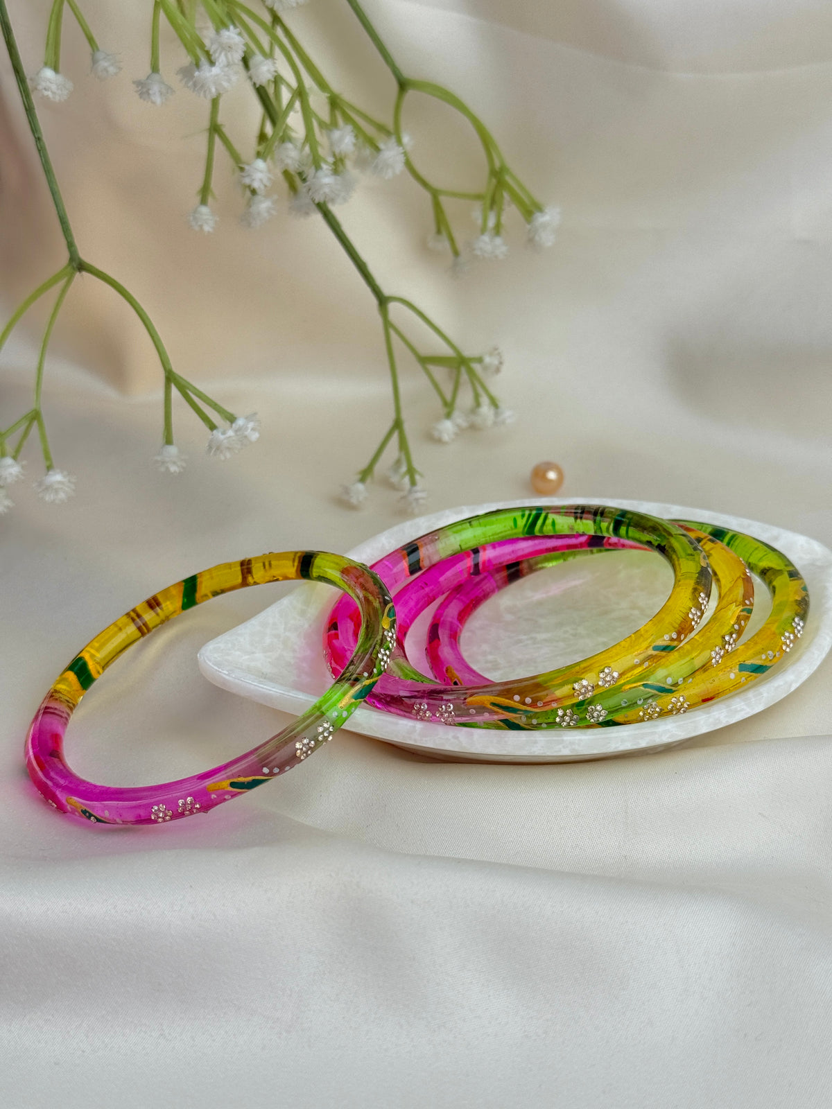 A set of colorful acrylic bangles with a marbled pattern of pink, green, yellow, and black, displayed on a small white dish with baby's breath flowers on a draped white satin background.