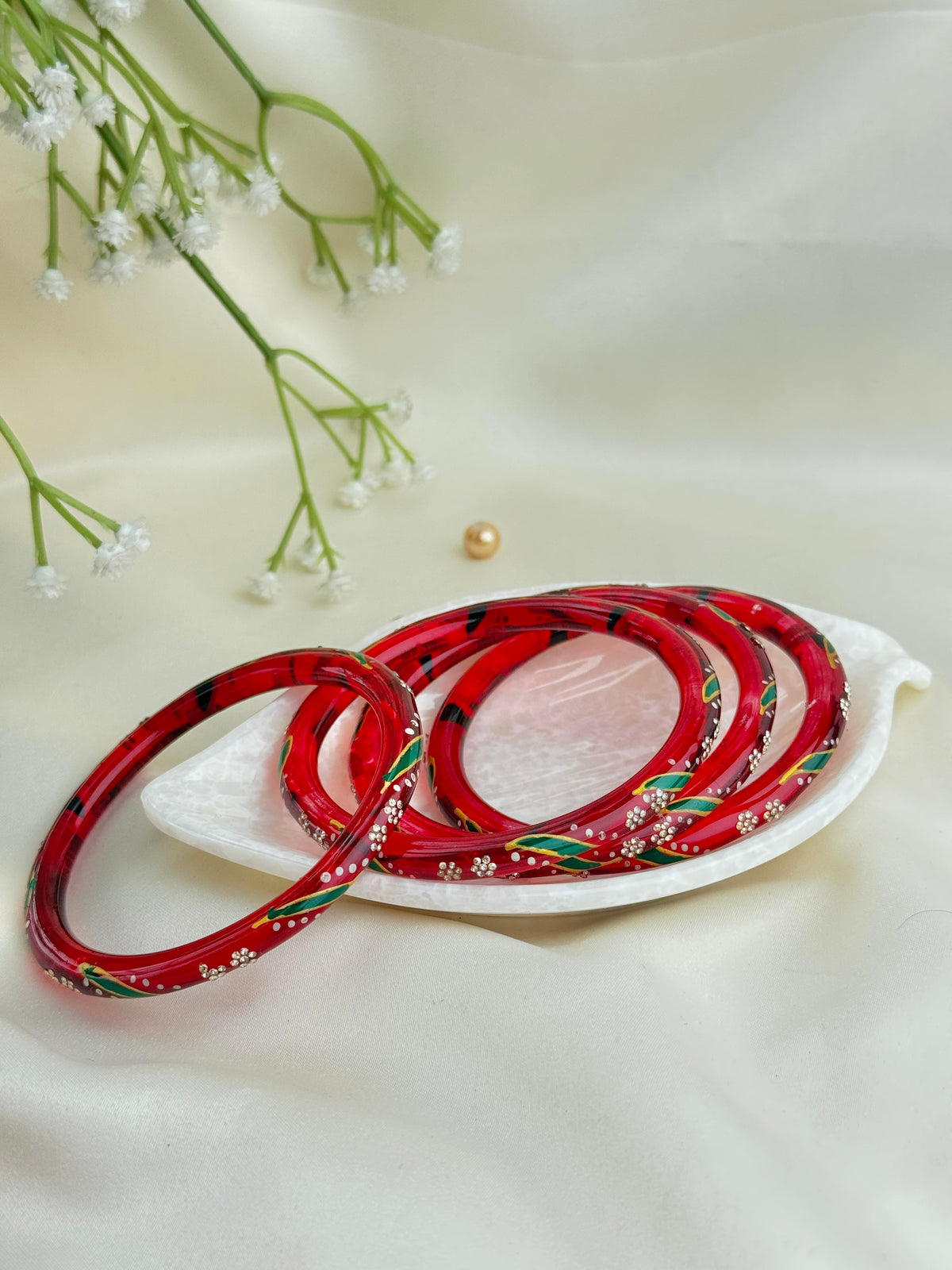A set of red bangles decorated with hand-painted green leaves and white floral motifs, arranged on a white ceramic dish with baby's breath flowers and a pearl in the background.