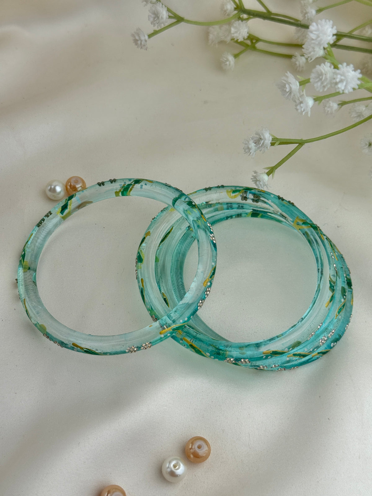 Two translucent aqua blue bangles with green and gold glittery patterns, displayed on a soft cream fabric background with decorative white flowers and small beads.