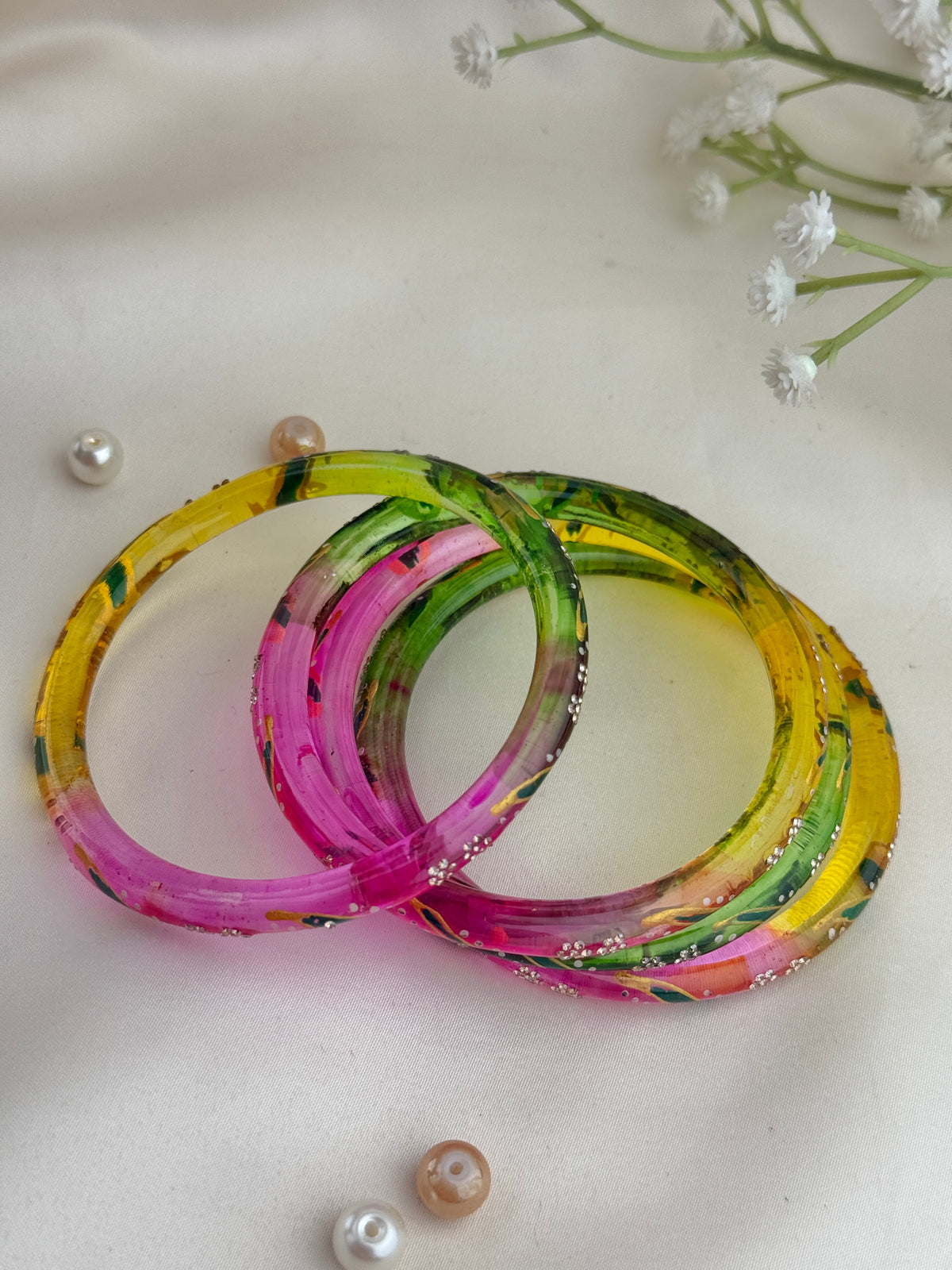 Three translucent acrylic bangles with a marbled paint pattern in yellow, pink, green, and orange, arranged on a light beige fabric background with decorative pearls and baby's breath flowers.
