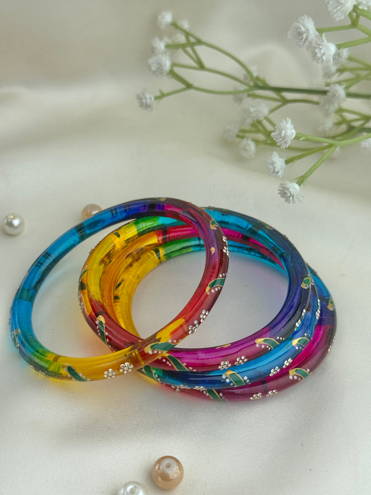 Four translucent acrylic bangles in a rainbow color pattern, decorated with small white floral inlays, arranged on a cream-colored fabric background with decorative white flowers and pearls.