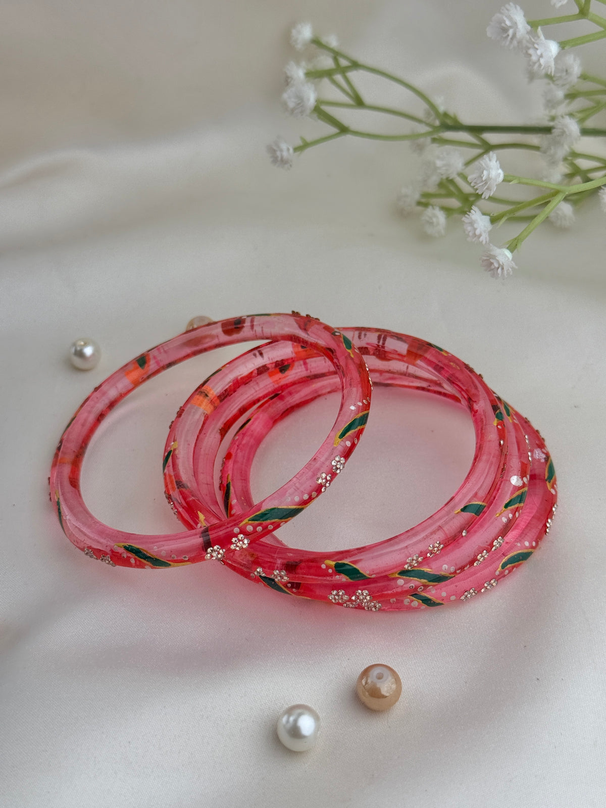Three pink acrylic bangles with floral and green leaf patterns, decorated with small silver rhinestones, arranged on a white fabric background with decorative pearls and baby's breath flowers.