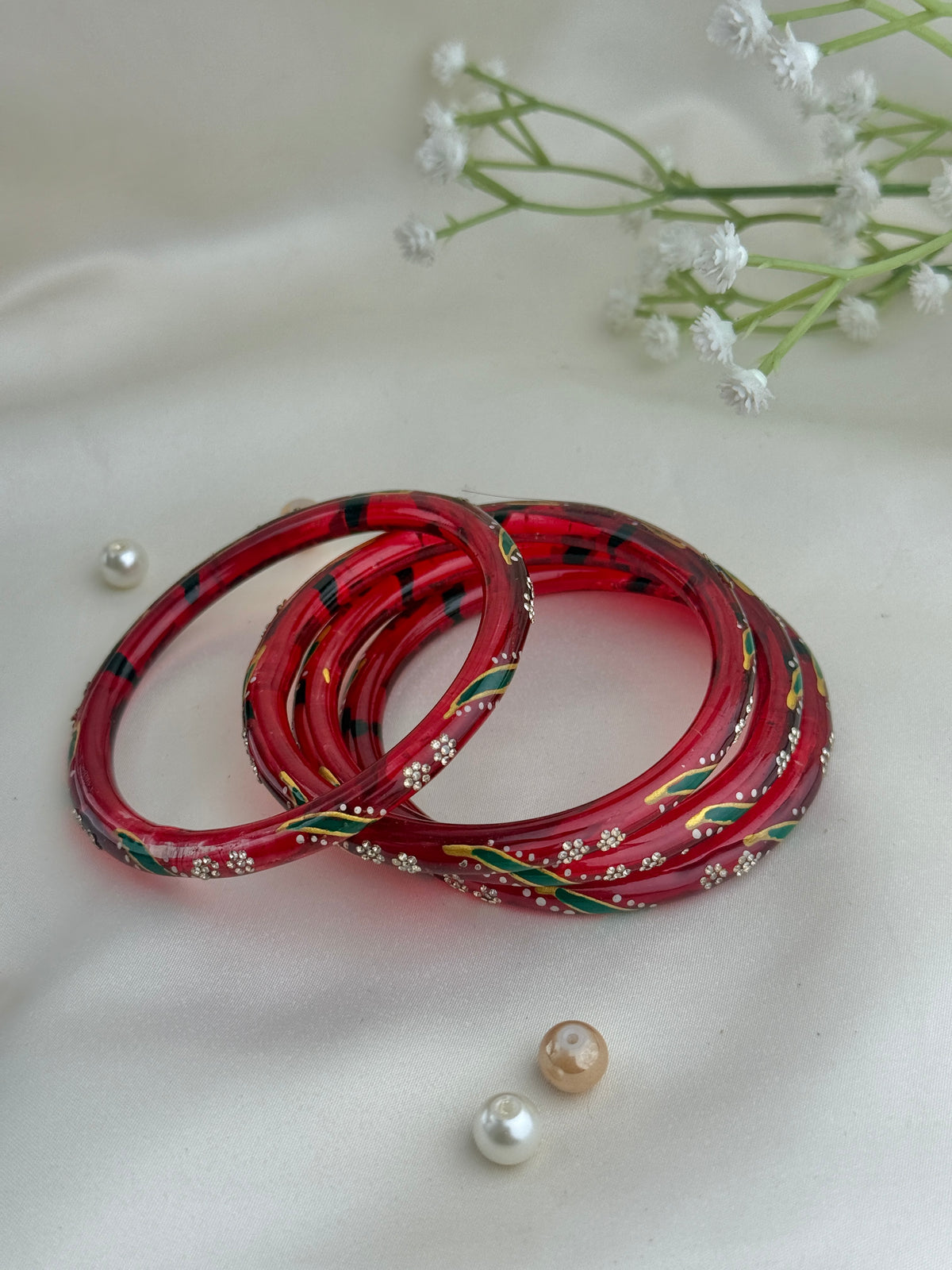 A set of red glass bangles decorated with black, green, and gold patterns and small white bead details, arranged on a white fabric background with decorative pearls and baby's breath flowers.