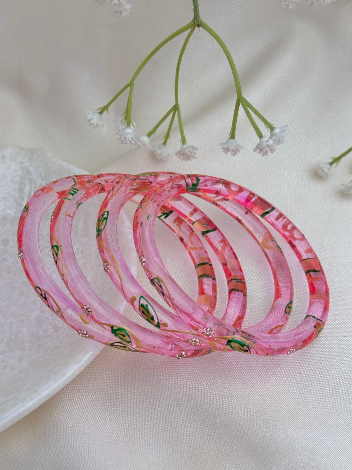 A set of five pink acrylic bangles with colorful floral patterns and small rhinestone accents, arranged on a white textured surface with delicate white flowers in the background.