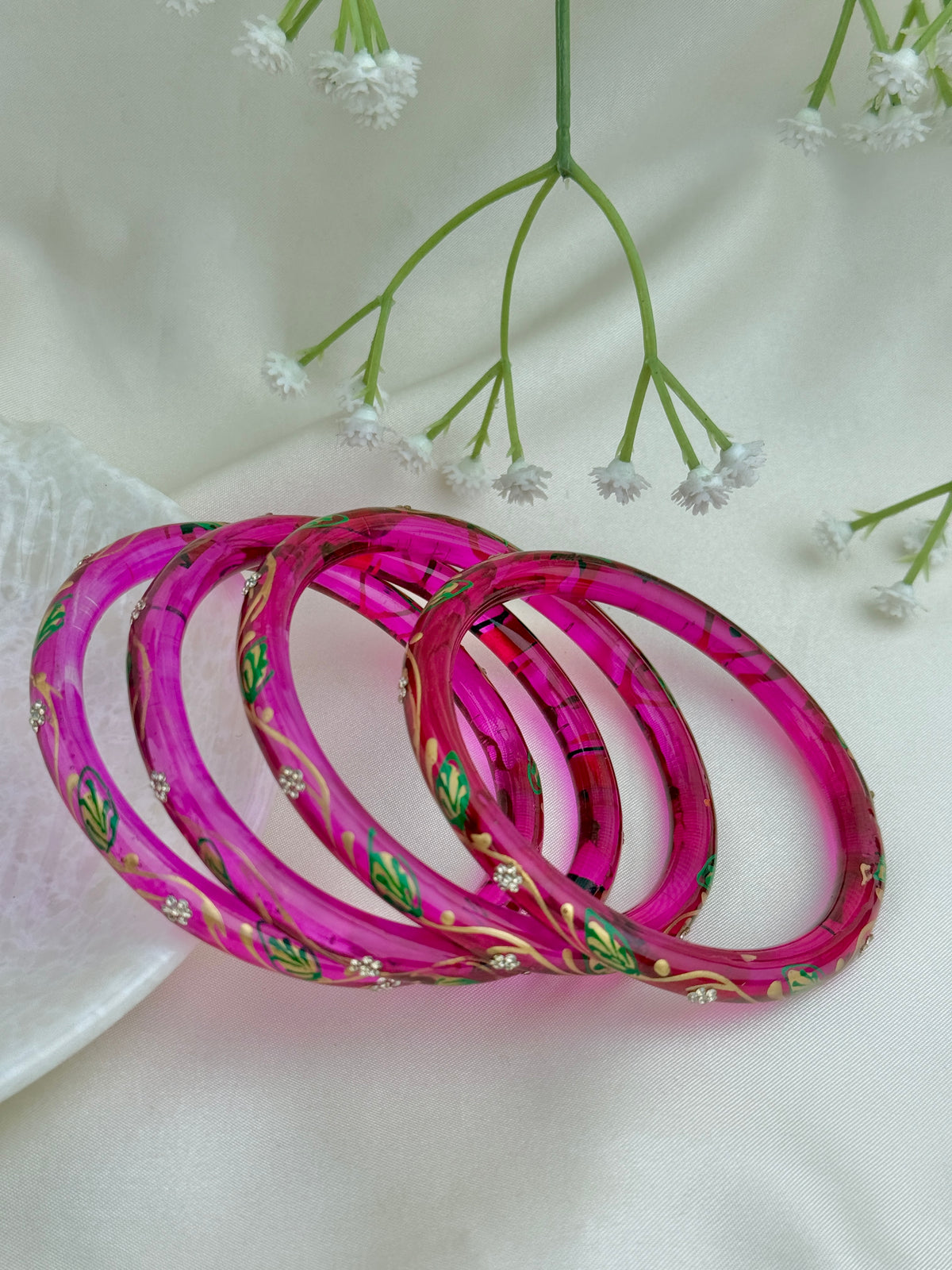 Four pink bangles with green and gold floral patterns, arranged on a white fabric background with small white flowers.