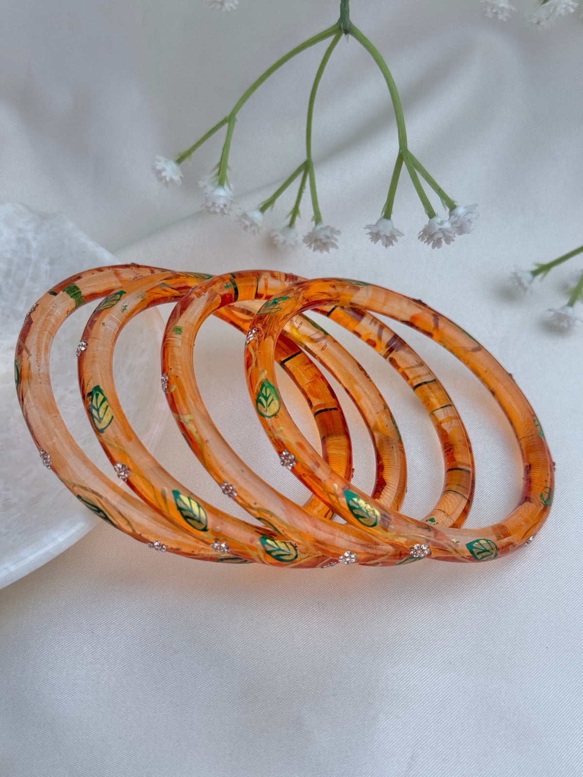 Four stacked peach-orange bangles with green leaf patterns and small silver rhinestone accents, displayed on a soft white fabric background with small white flowers.