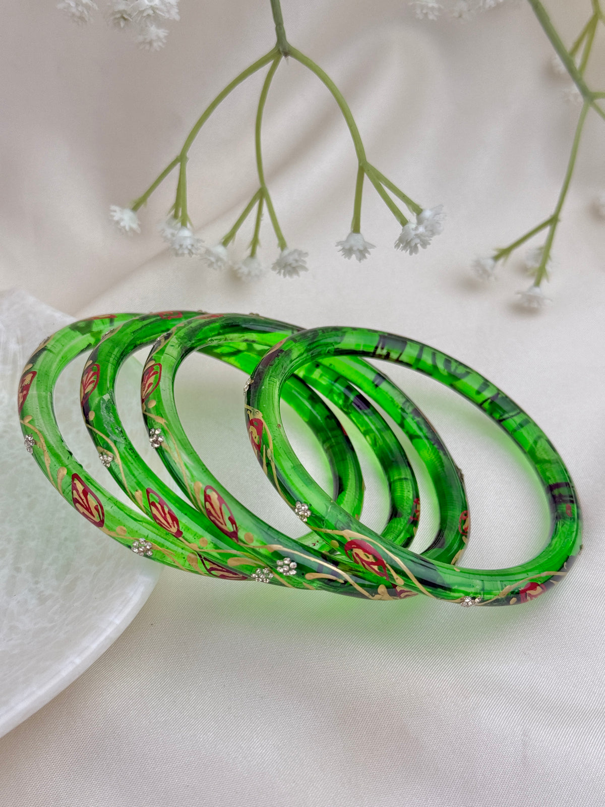Four green acrylic bangles with red floral patterns and small silver rhinestone accents, arranged on a white fabric background with baby's breath flowers.