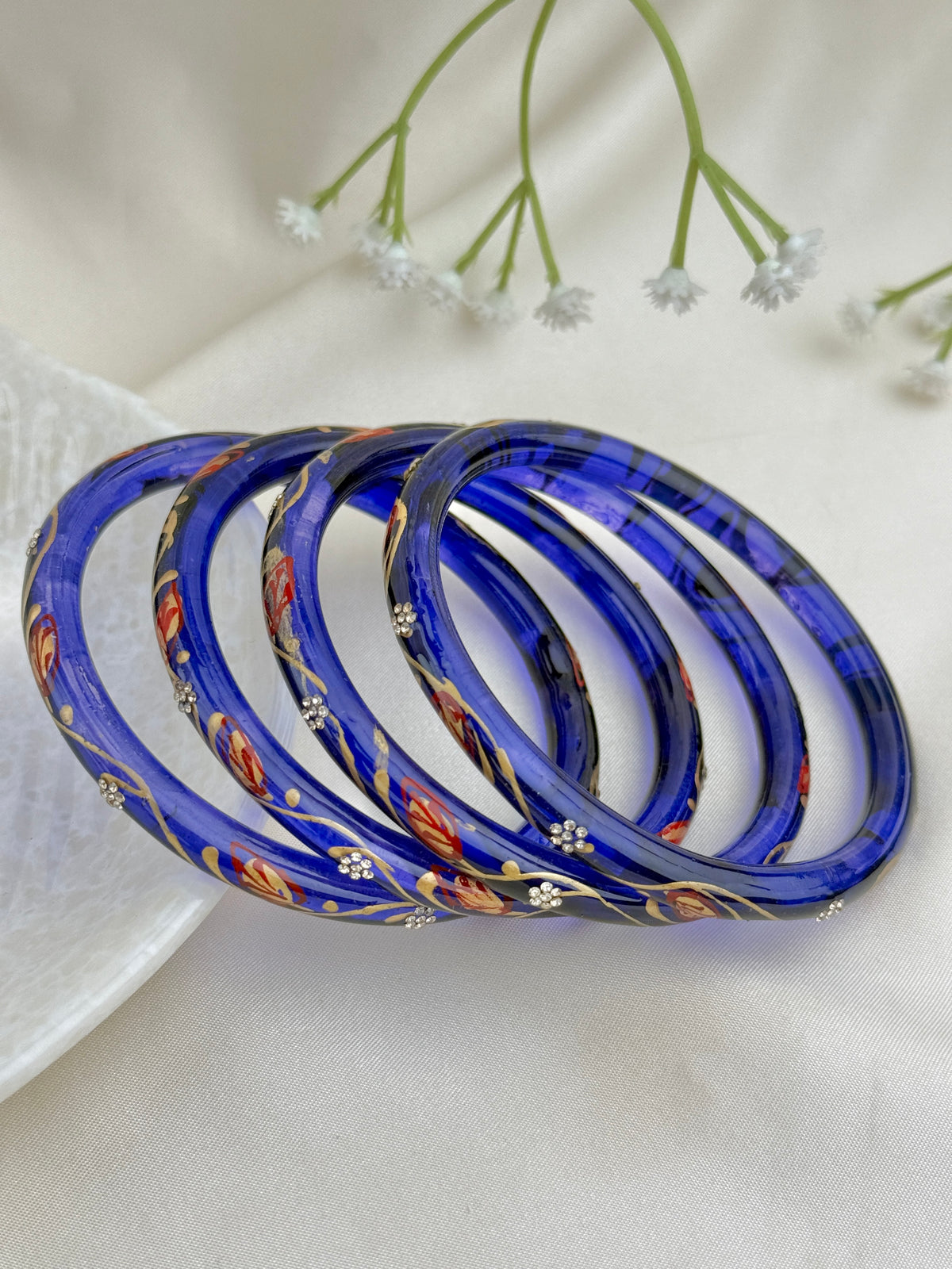 Four blue glass bangles with red, gold, and white floral patterns, arranged on a white fabric background with small white flowers.