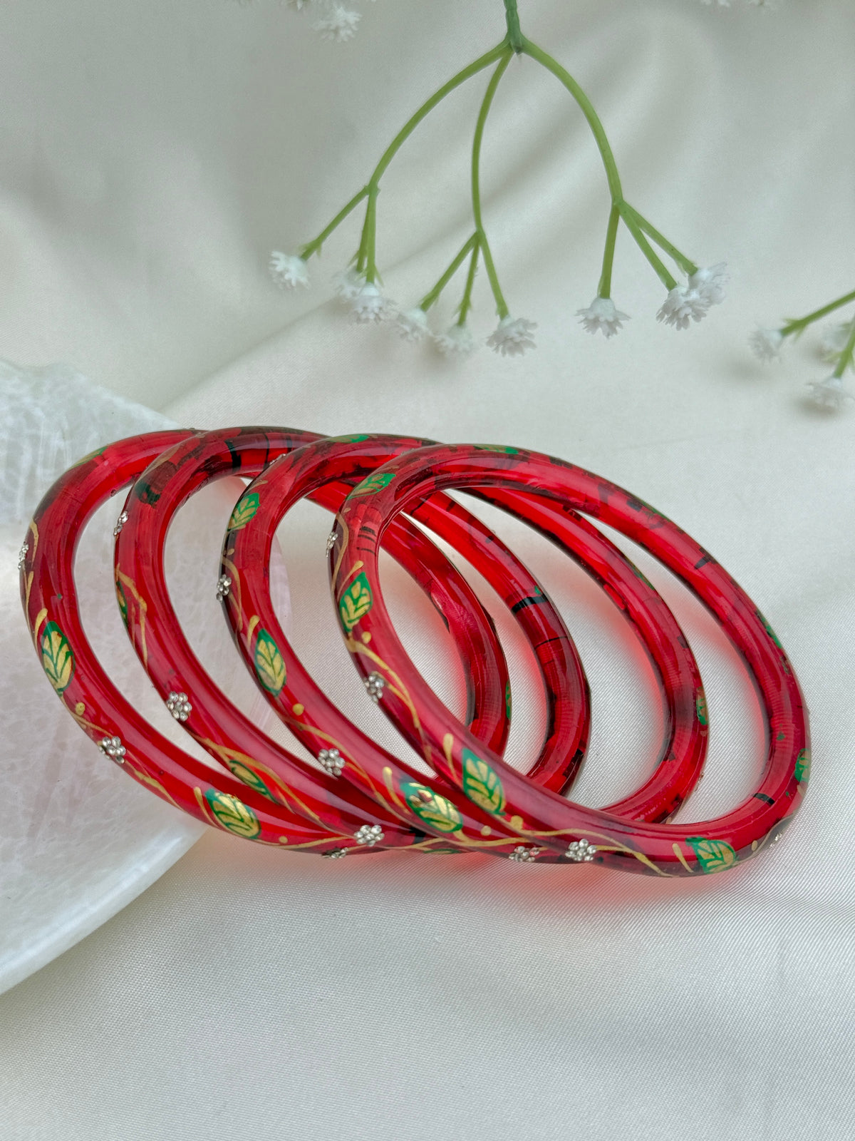 Four red bangles with green leaf and gold floral patterns, arranged in a fan shape on a white fabric background with small white flowers.