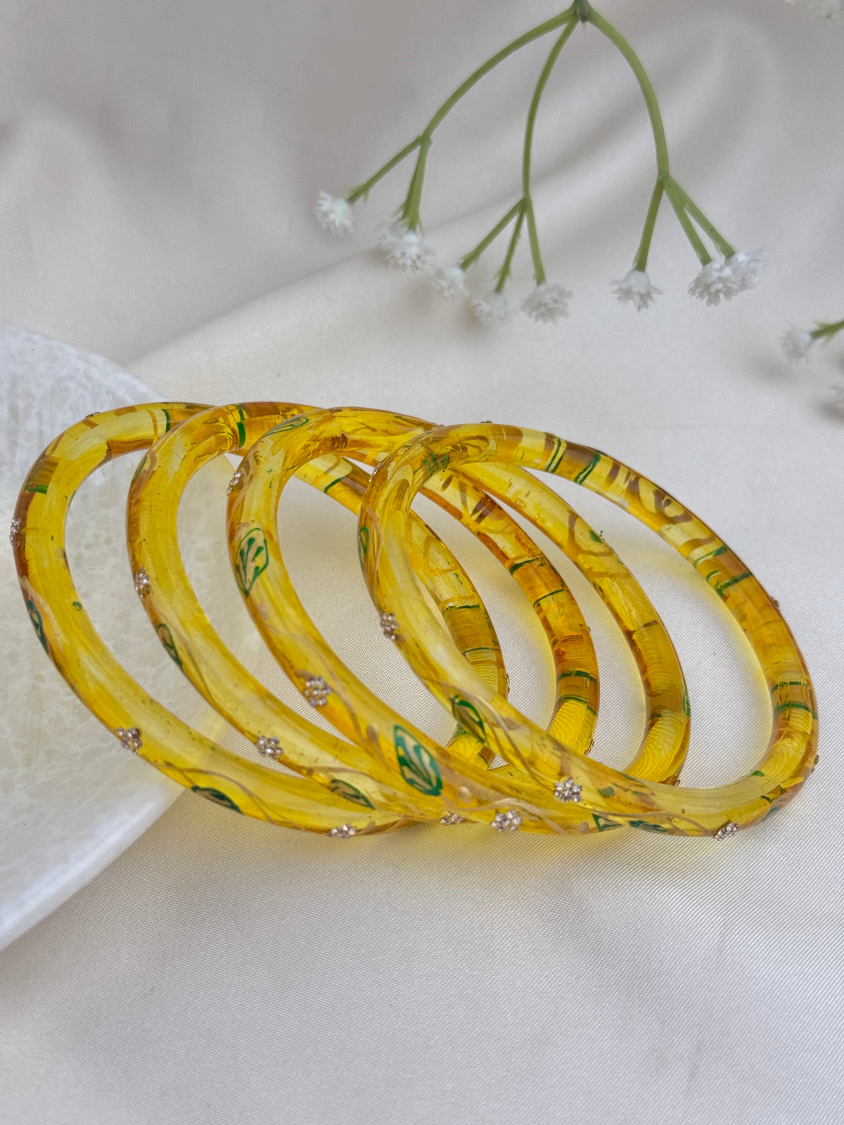 Four yellow translucent bangles with green leaf patterns and small clear rhinestone accents, arranged on a soft white fabric background with small white flowers.