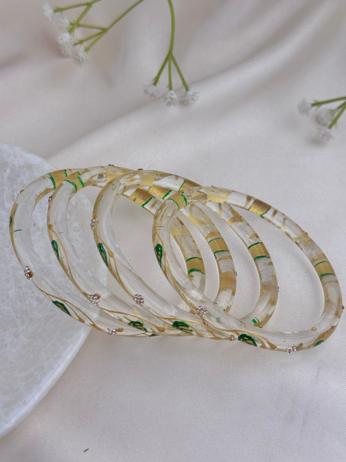 A set of five stacked translucent resin bangles with a gold and green leaf pattern, resting on a white fabric background with small white flowers.