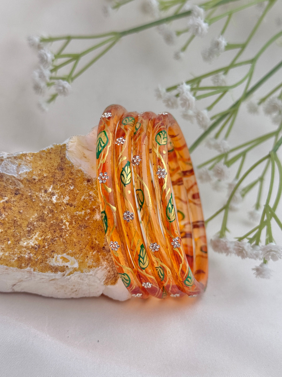 A set of translucent orange bangles decorated with green leaf patterns and small silver rhinestone accents, displayed on a textured stone against a soft white background with delicate white flowers.