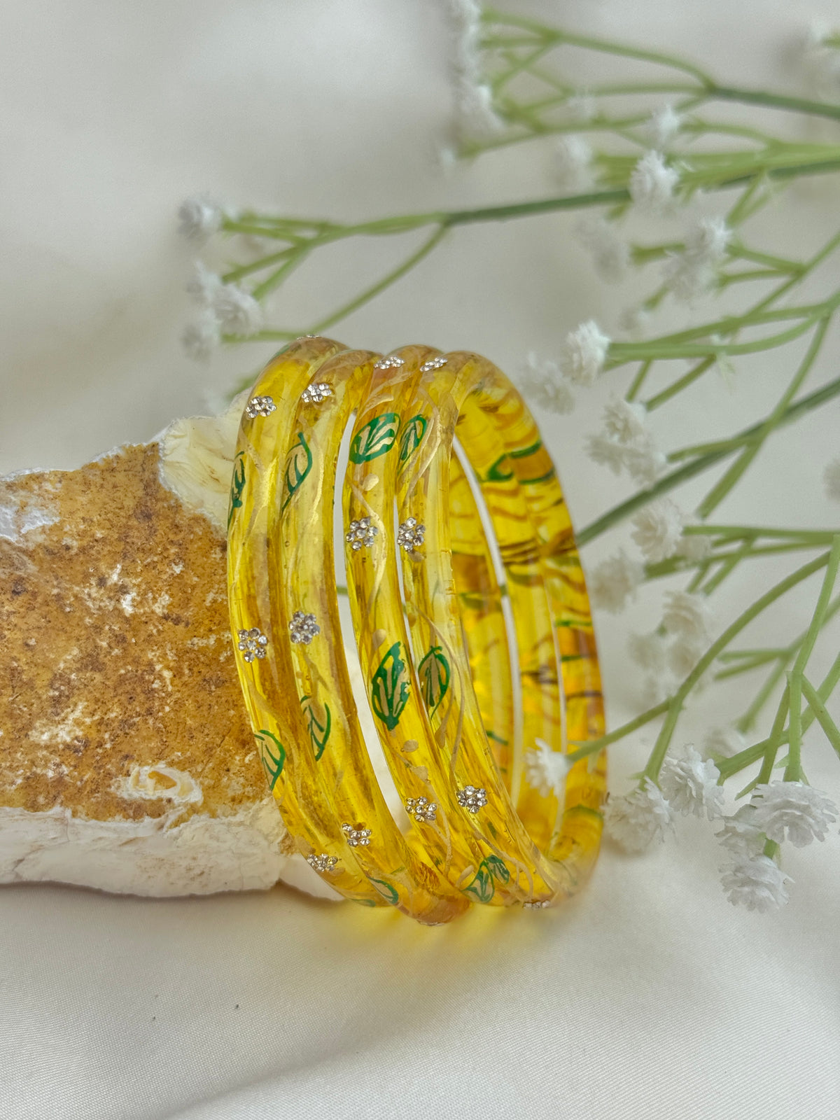A set of translucent yellow bangles with green leaf patterns and small silver rhinestone accents, displayed on a textured stone against a soft cream fabric backdrop with white baby's breath flowers.