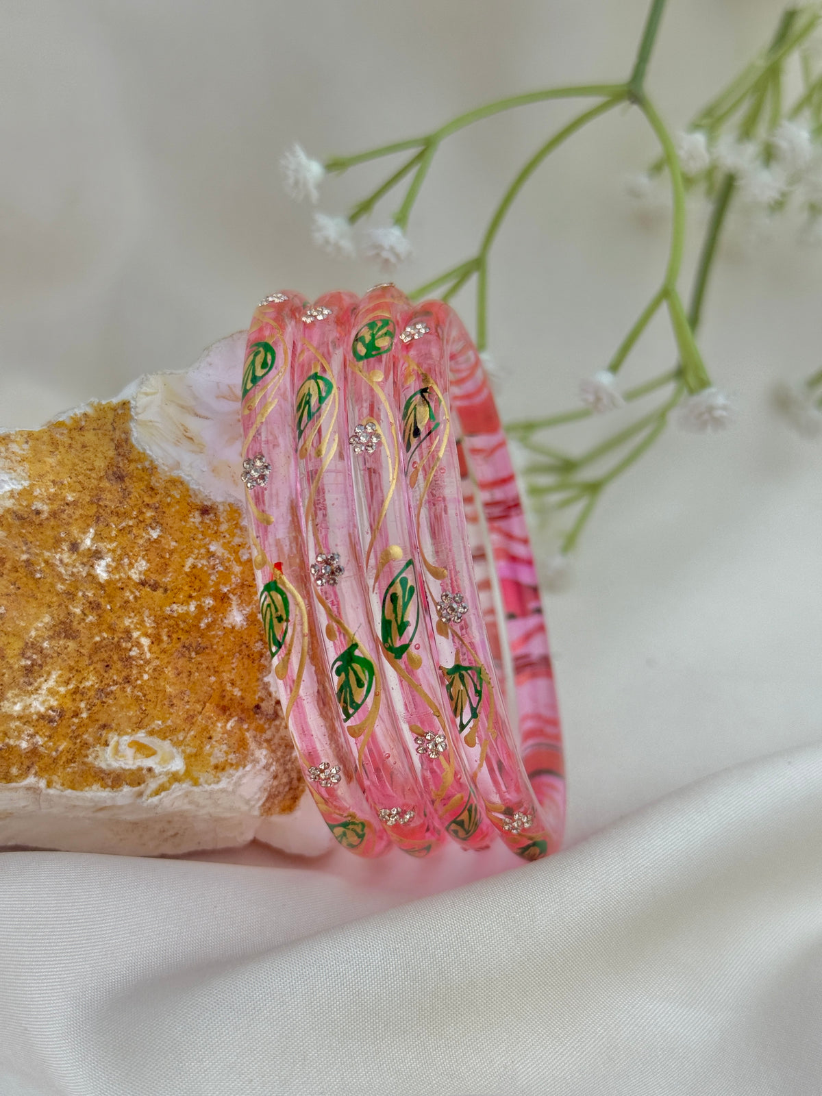 A set of four pink glass bangles with green leaf patterns and small gold and silver accents, displayed on a textured stone next to white satin fabric and baby's breath flowers.