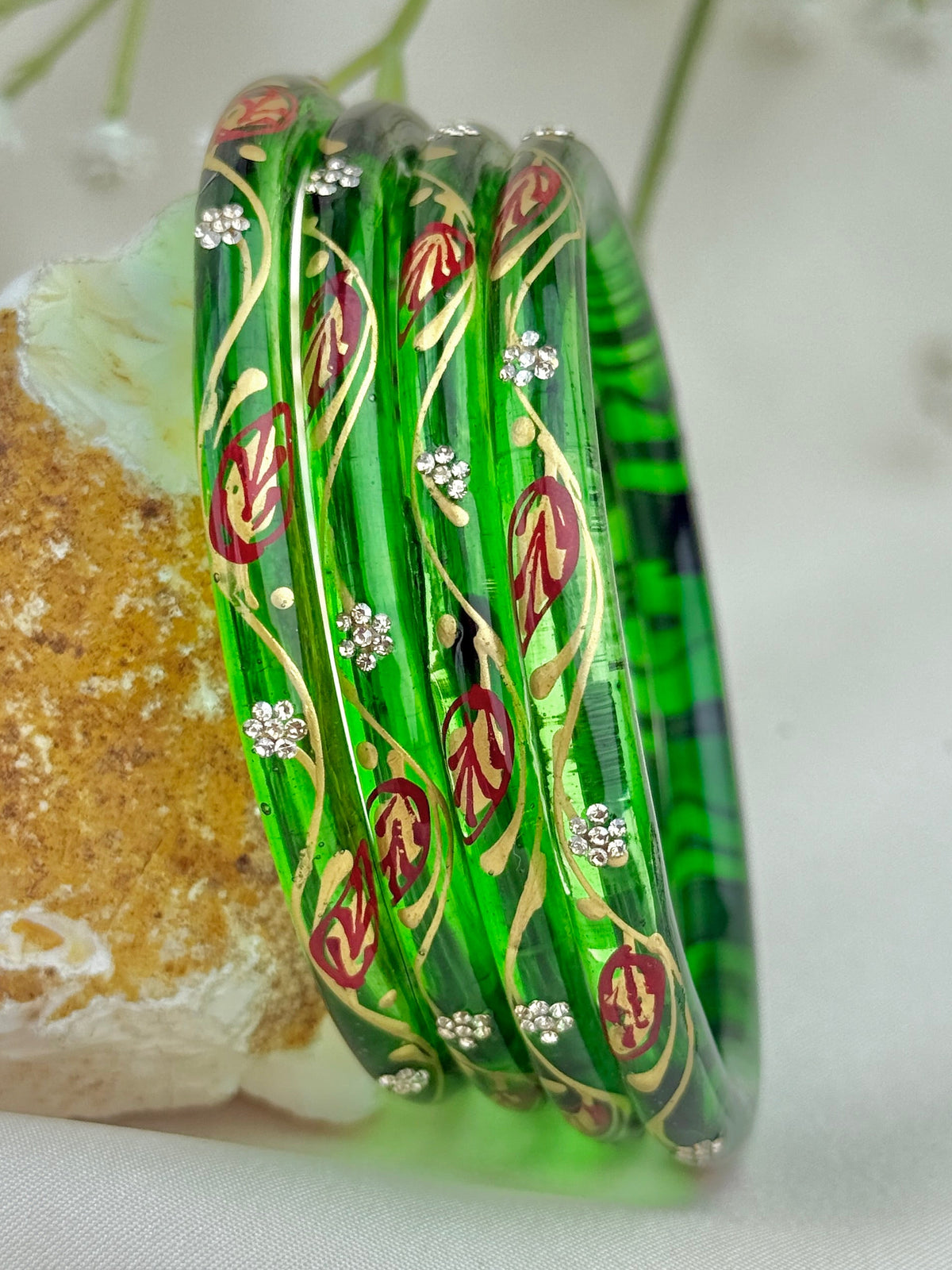 A set of green glass bangles with hand-painted red and gold leaf patterns and small white floral details, displayed on a textured stone against a soft cream fabric background with baby's breath flowers.