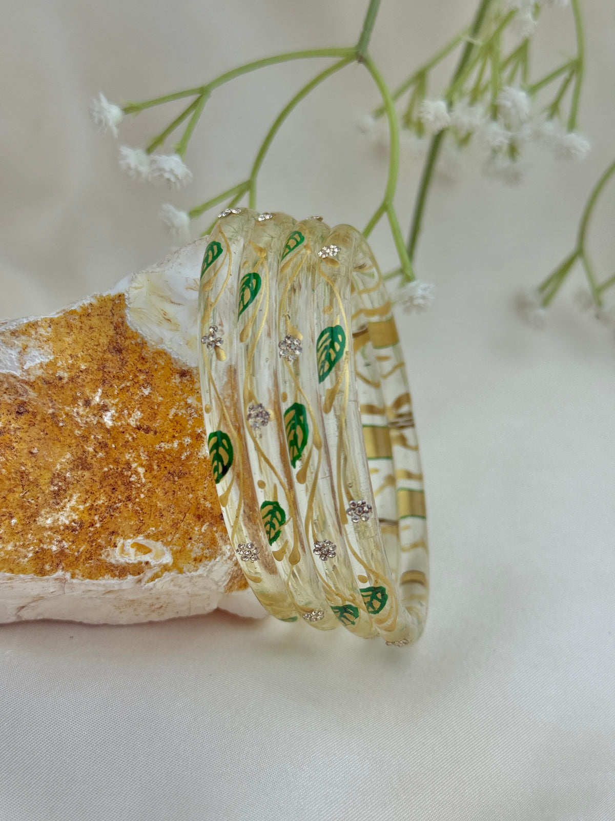 A set of five transparent glass bangles with green leaf patterns and small silver rhinestone accents, displayed on a textured orange and white stone with delicate white flowers in the background.