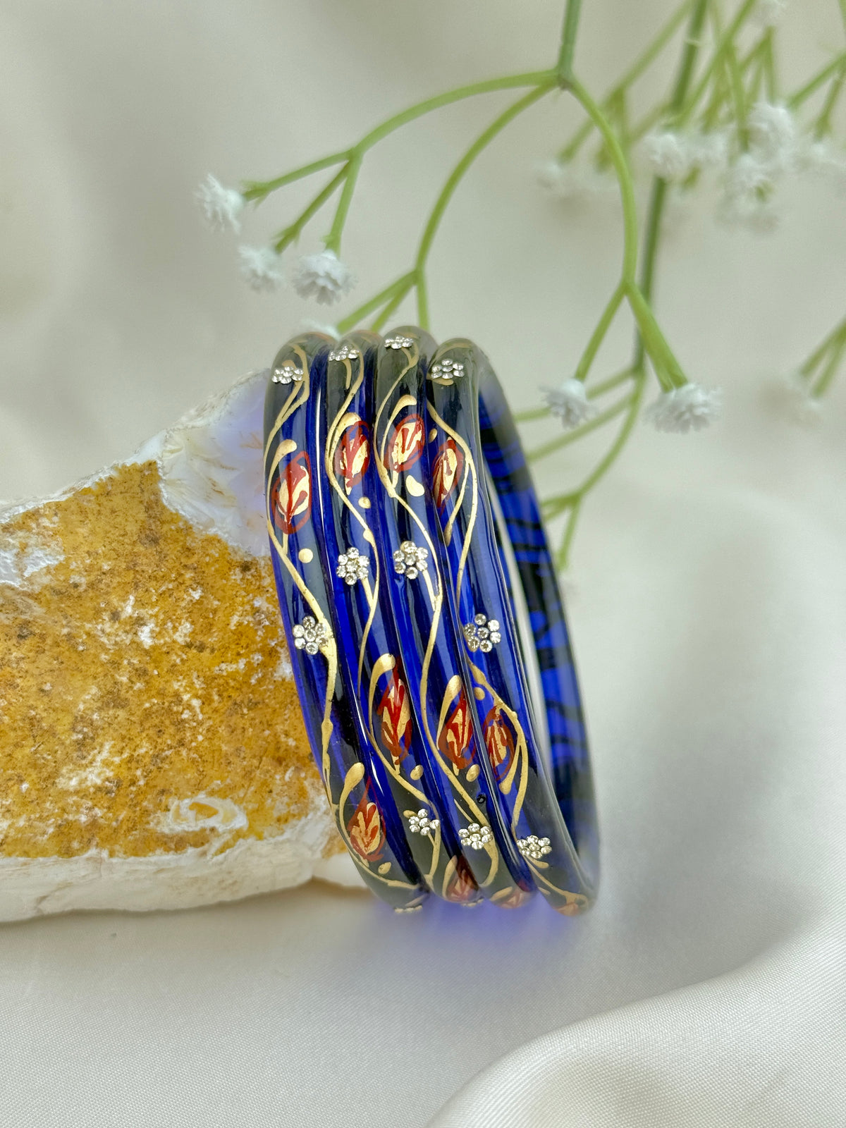 Four blue glass bangles with gold and red floral patterns, displayed on a gold-colored rock against a soft white fabric background with small white flowers.