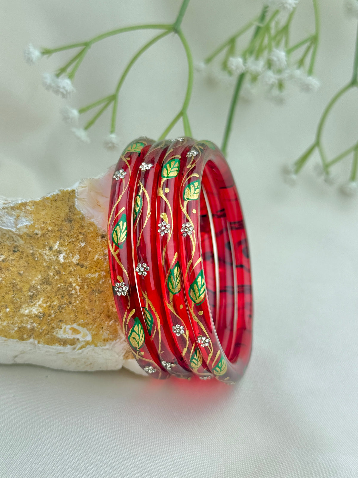 A set of five red bangles with hand-painted green leaves, gold vines, and small white floral motifs, displayed on a textured stone with white baby's breath flowers in the background.