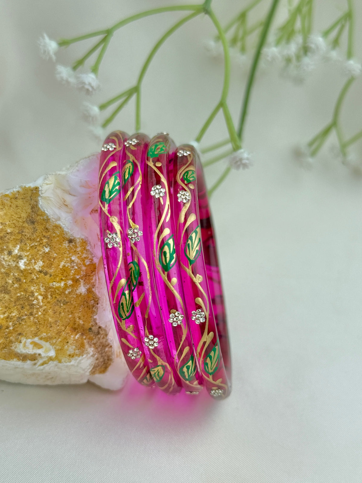 A set of five glossy pink bangles decorated with gold leaf patterns, green floral motifs, and small rhinestone clusters, displayed on a white surface next to a textured stone.