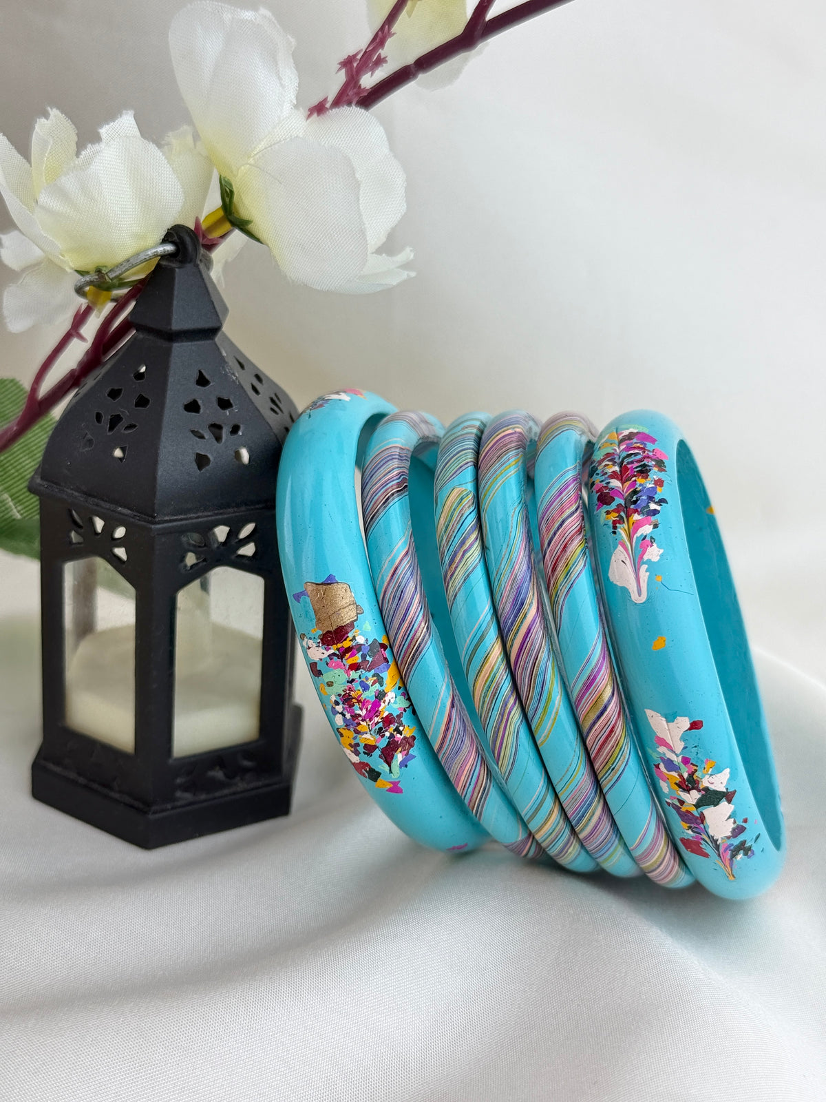 A set of turquoise bangles with colorful floral and striped patterns, arranged next to a black decorative lantern with white flowers on a white fabric background.