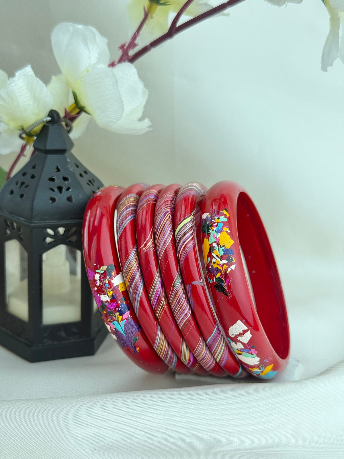 A set of red bangles with colorful floral and striped patterns, arranged on a white fabric surface next to a black decorative lantern with white flowers in the background.