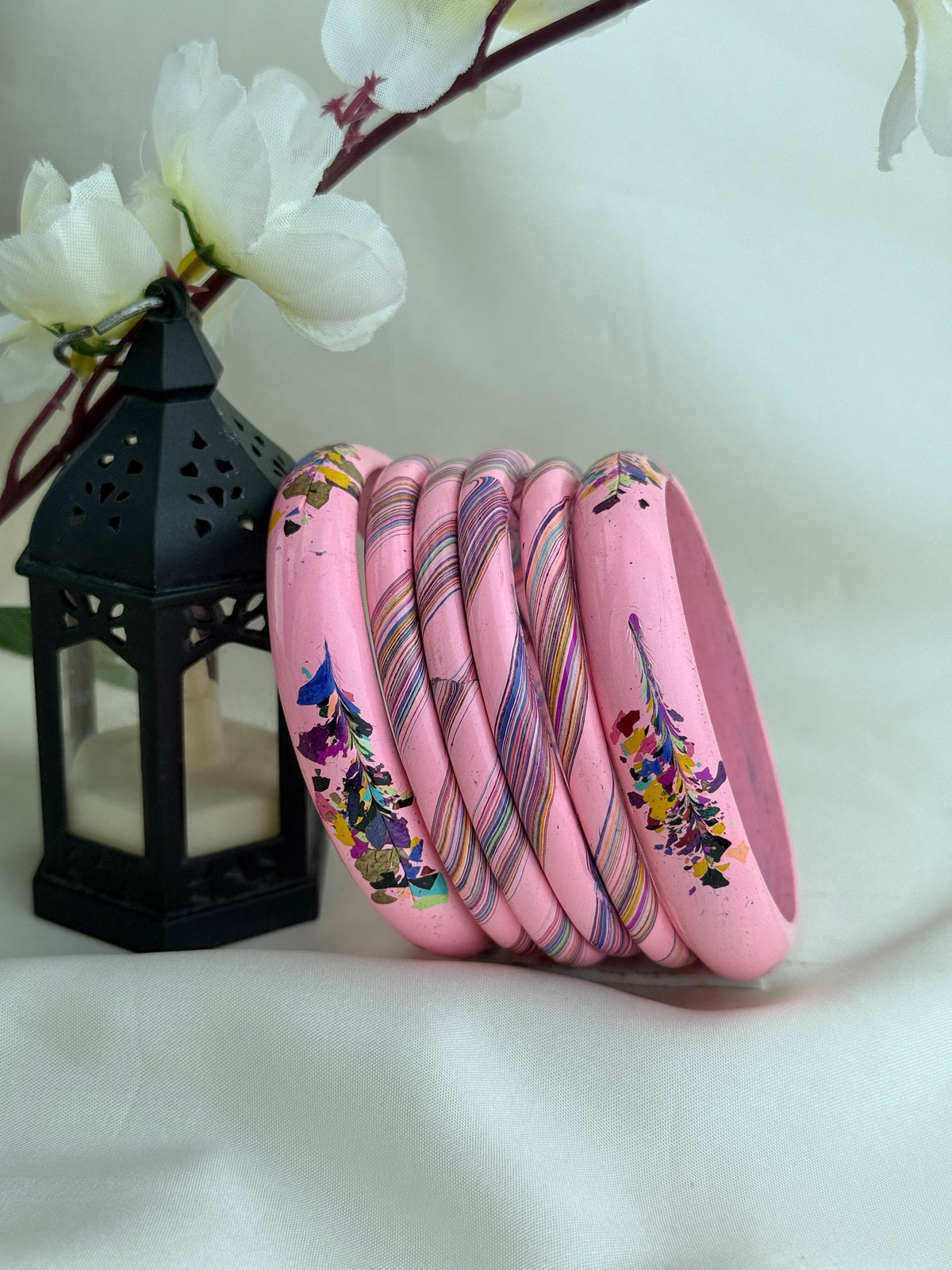 A stack of pink bangles with colorful floral and striped patterns, displayed next to a black decorative lantern with white flowers in the background.