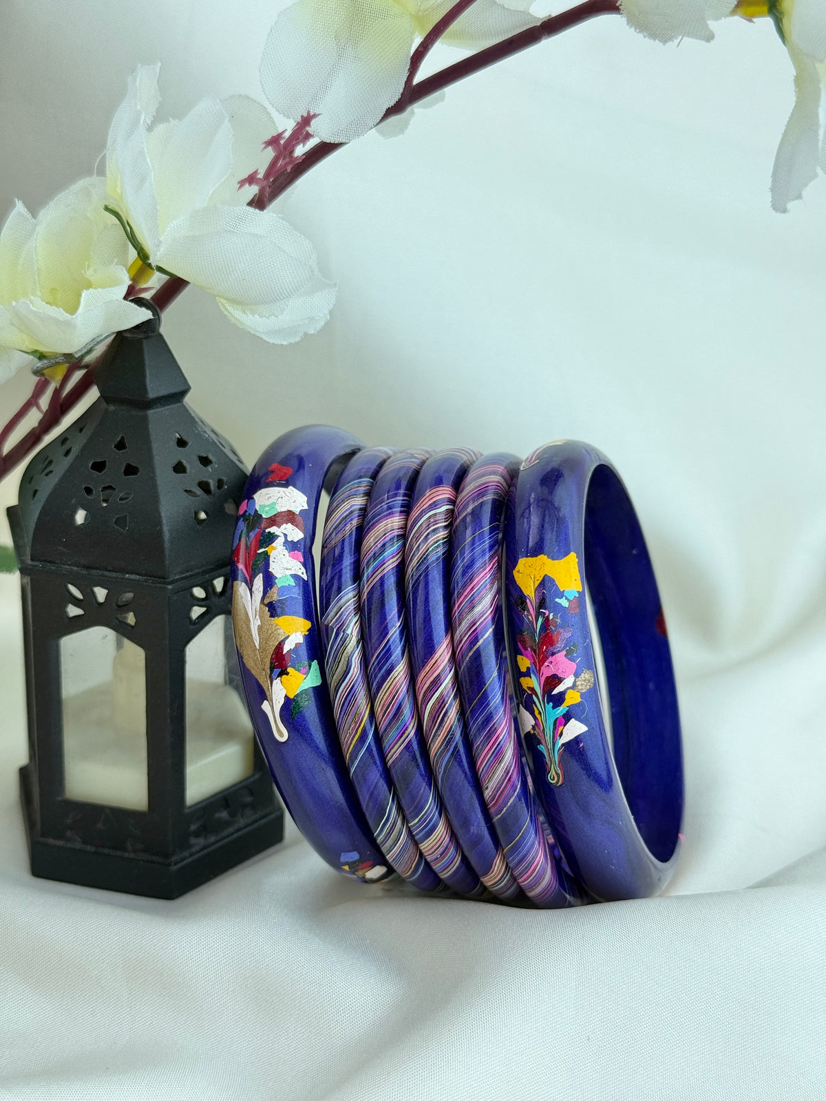 A set of nine traditional Indian copper bangles with colorful hand-painted floral and geometric designs, arranged next to a black decorative lantern on a draped white fabric background.