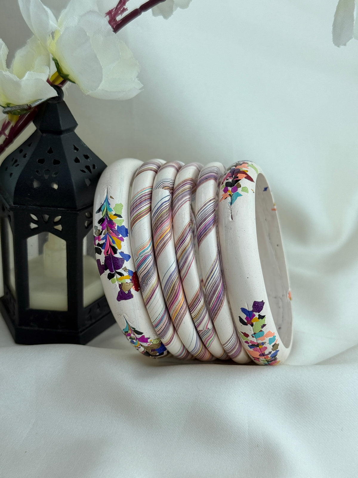 A set of nine stacked bangles, including some with colorful floral prints and others with diagonal marbled stripes in pink, purple, and gold, displayed on a white fabric backdrop next to a black lantern.