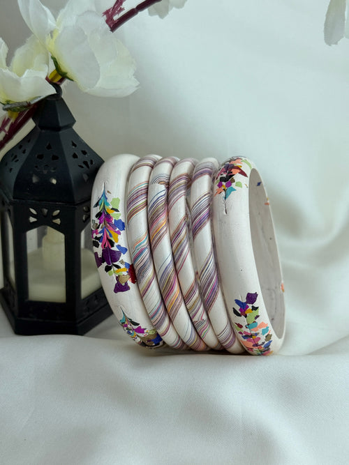 A set of nine stacked bangles, including some with colorful floral prints and others with diagonal marbled stripes in pink, purple, and gold, displayed on a white fabric backdrop next to a black lantern.