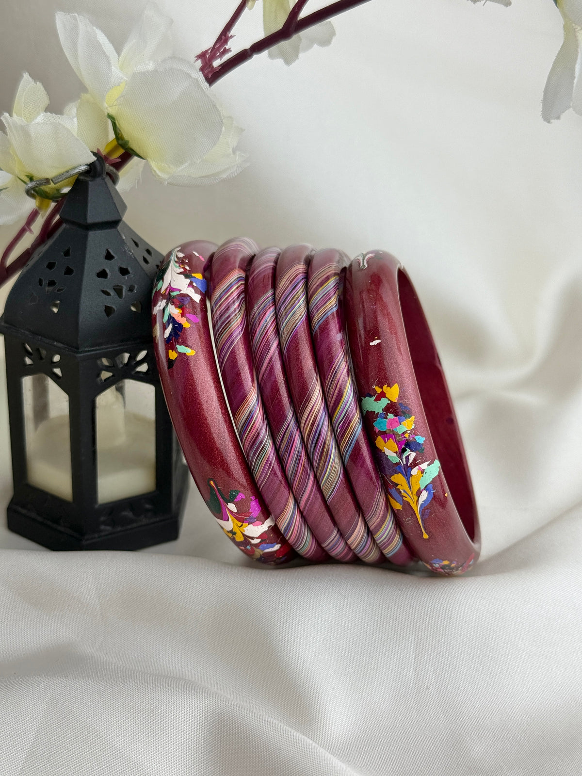 A set of nine stacked wooden bangles with a deep red base, featuring hand-painted floral motifs and striped patterns in pink, yellow, blue, and green, displayed next to a black lantern on a white fabric background.