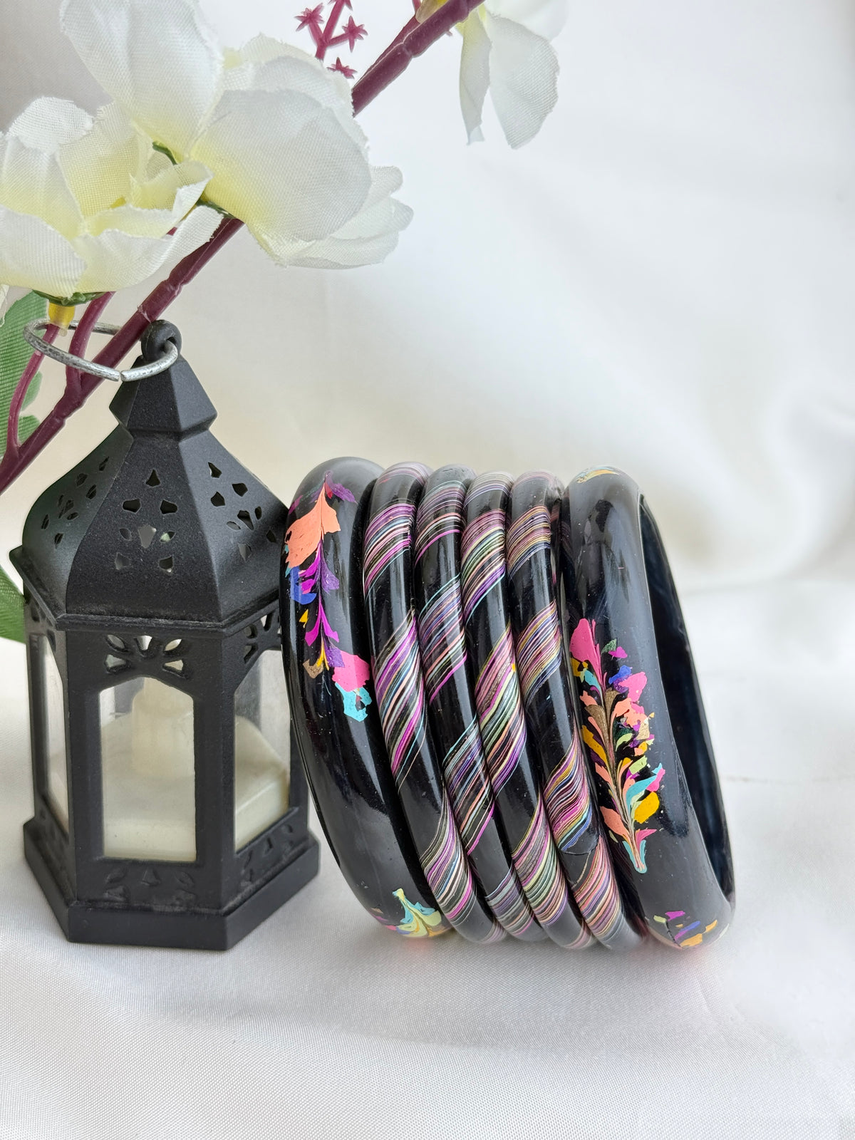 A set of nine black bangles with colorful floral and striped patterns, displayed next to a black decorative lantern with white flowers in the background.