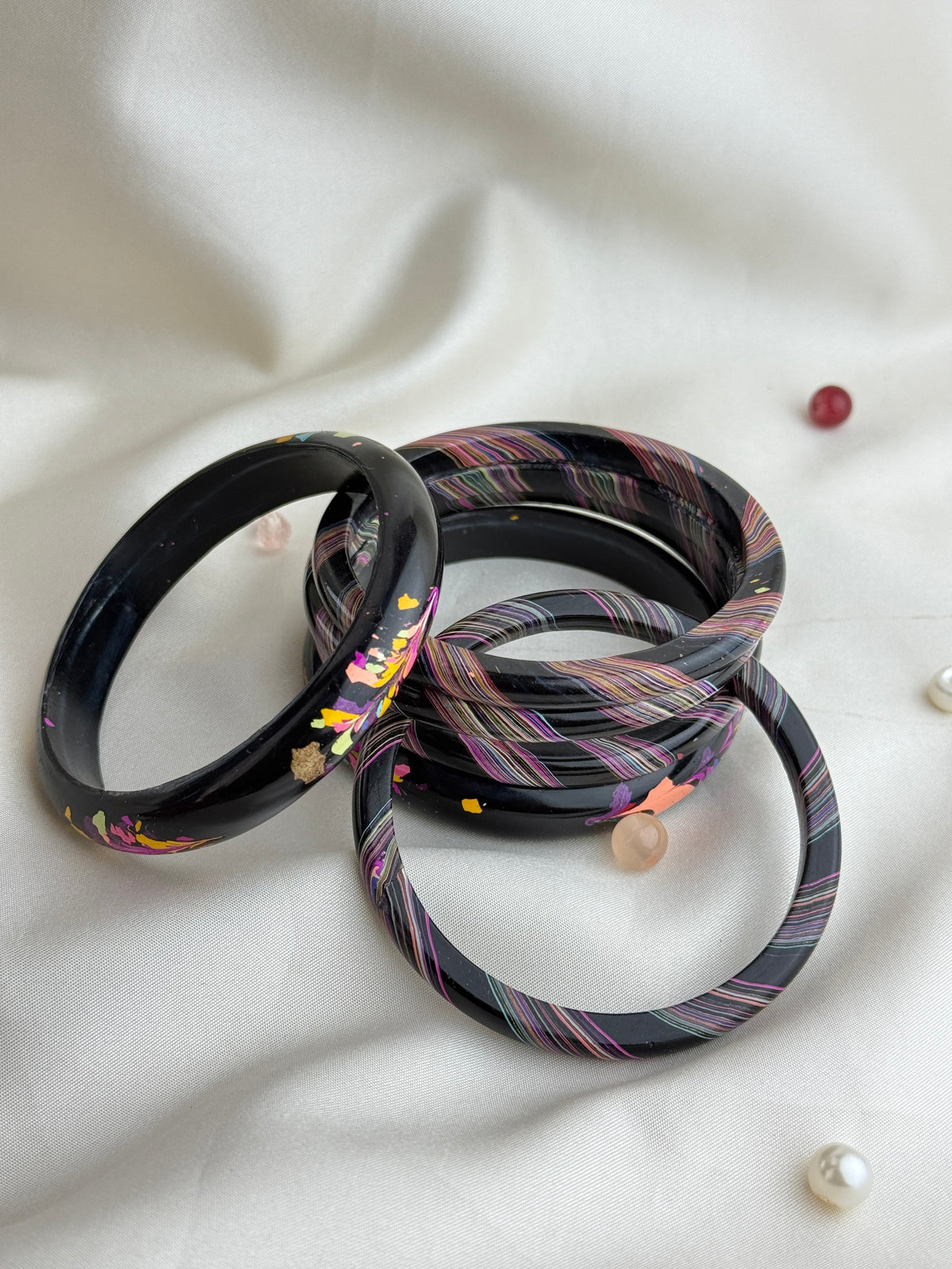 Three bangle bracelets arranged on a white fabric background. Two bracelets are solid glossy black with small multicolor floral inclusions, and one bracelet has a black base with thin pink, purple, and orange striped patterns.
