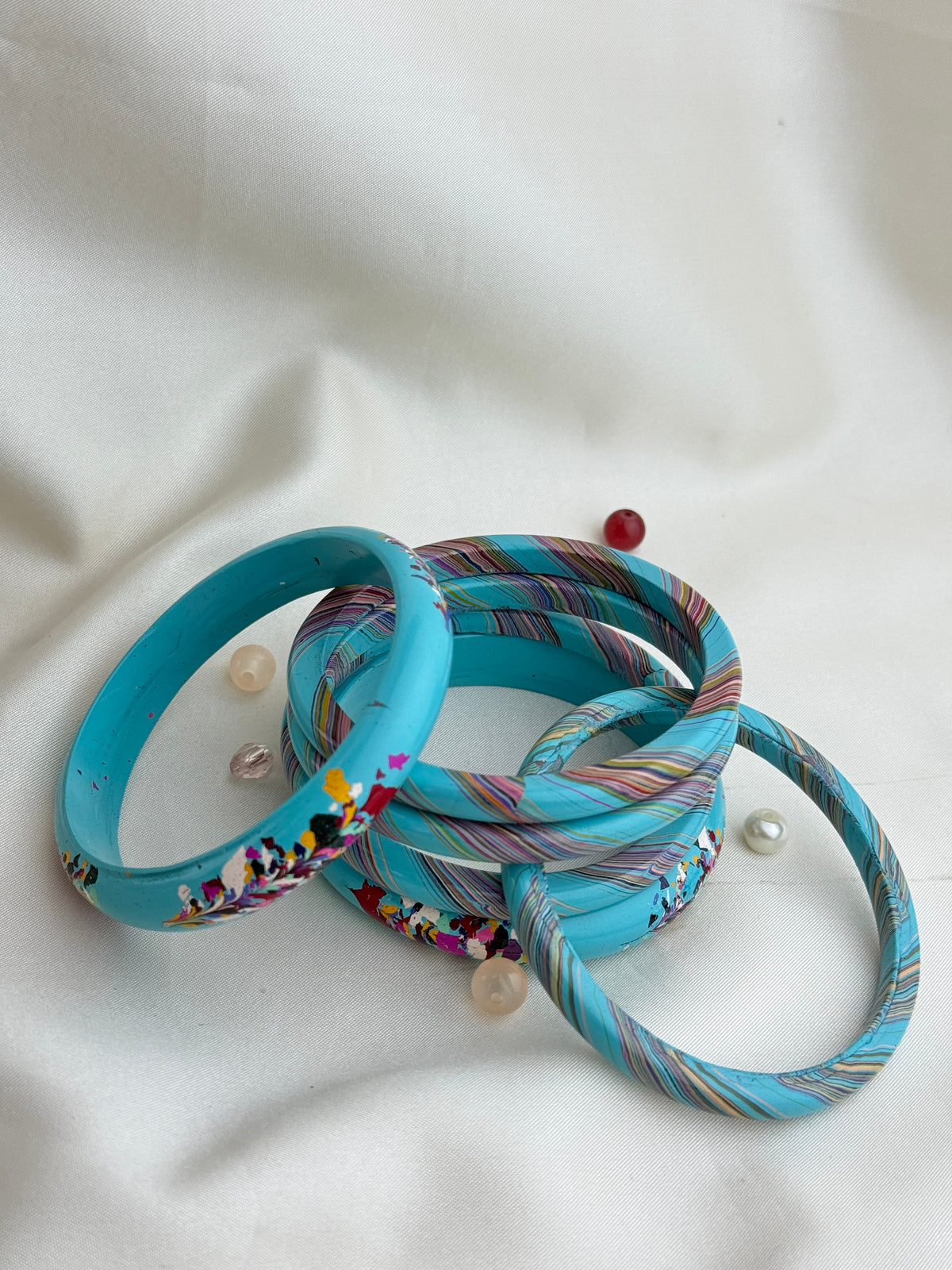 A set of turquoise bangles with colorful printed and striped patterns, arranged on a white fabric background.