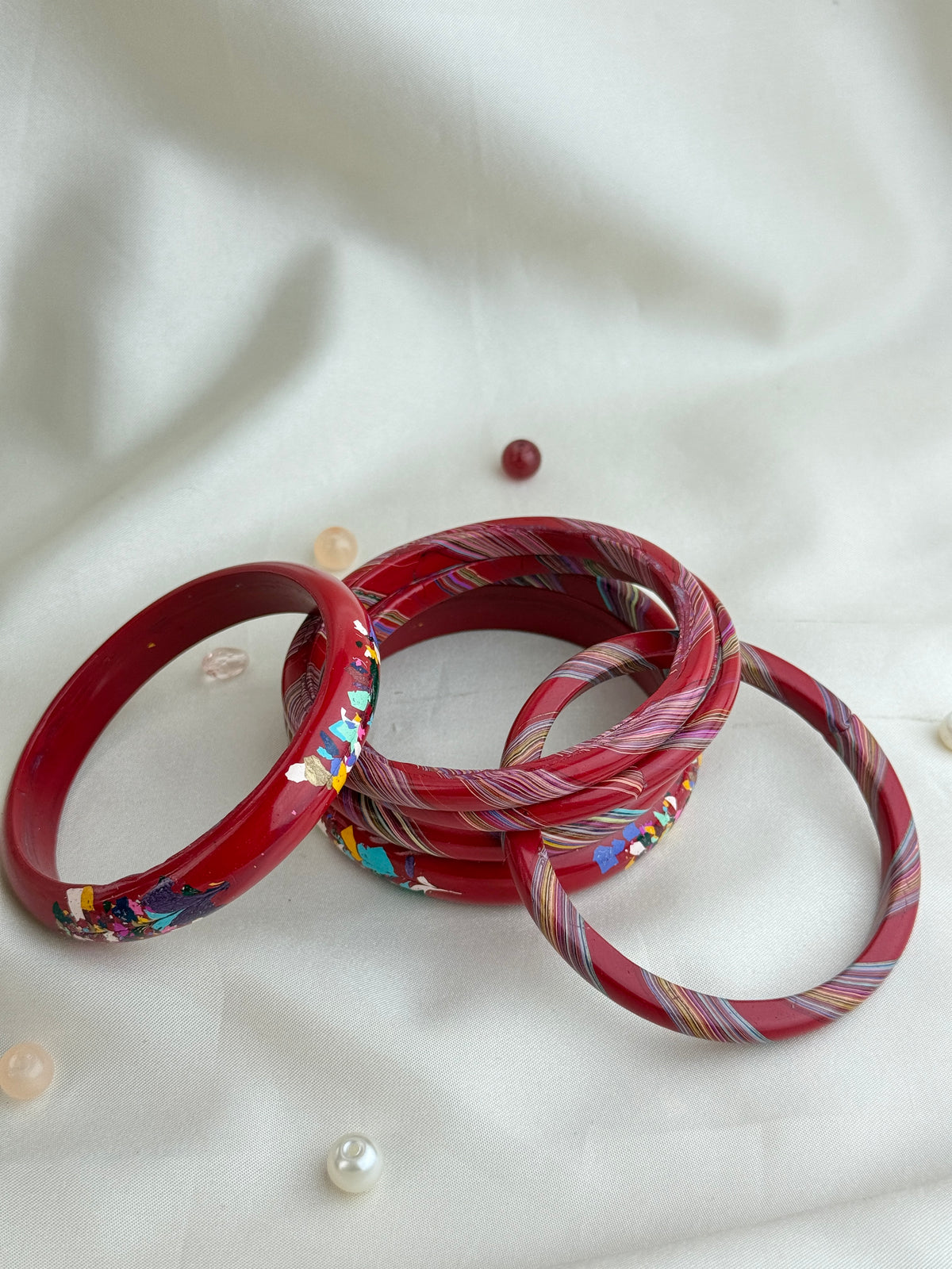 A set of red bangles, including one solid red bangle and several striped red, pink, and white bangles with colorful inlay details, arranged on a white fabric background.
