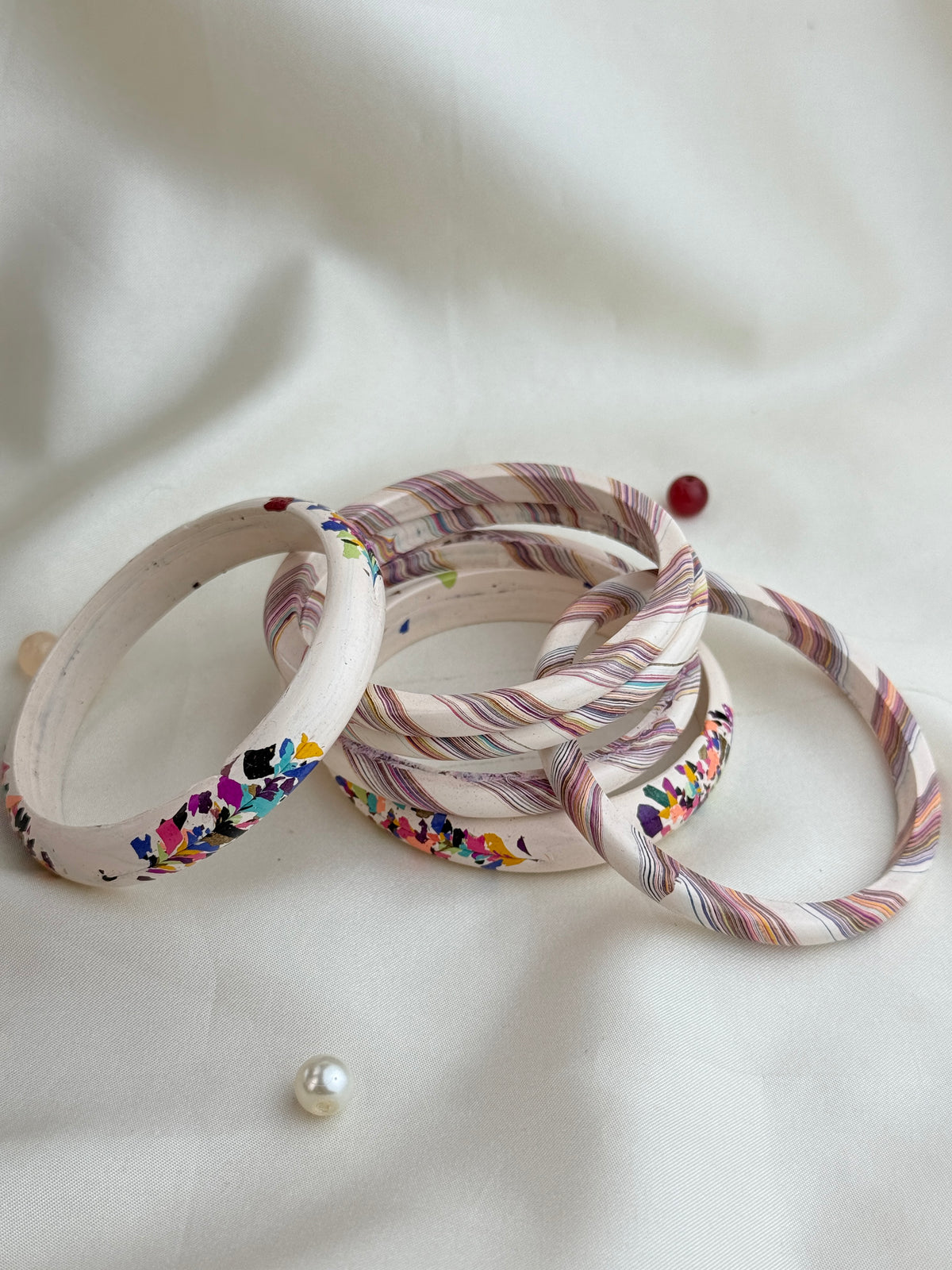 A set of handmade bangles made from wrapped wooden dowels, featuring colorful floral patterns and striped designs in pink, purple, blue, and green on a cream base, displayed on a soft white fabric background.