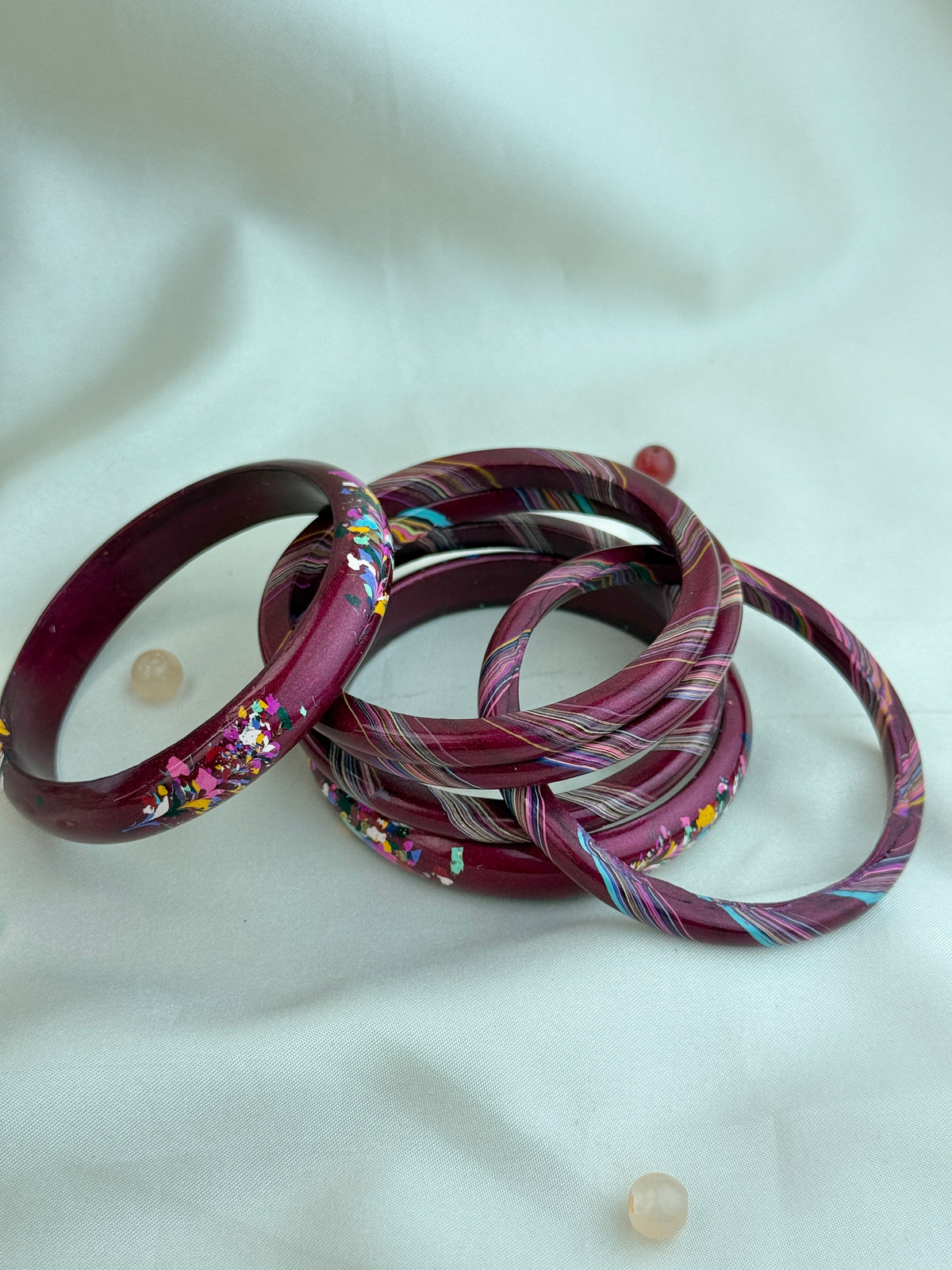 A set of five marbled acrylic bangles in deep burgundy and purple tones, some with embedded glitter and others with diagonal multicolor swirls, displayed on a white fabric background.