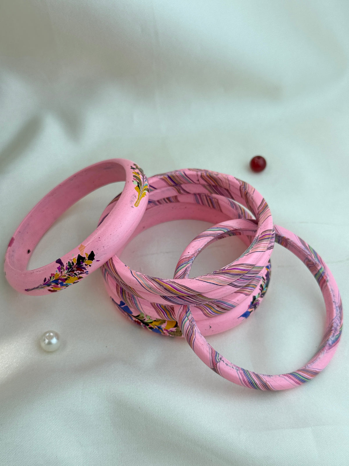 A set of handmade pink bangles, including one solid pink bangle with colorful floral patterns and several others with pink and blue striped woven fabric, displayed on a white fabric background.